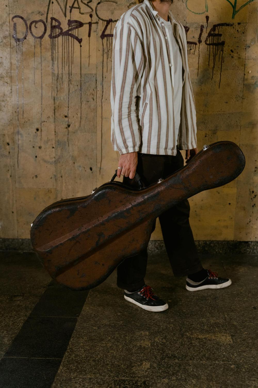 A person wearing a striped shirt and black pants holds a worn guitar case, standing against a graffiti-covered wall.