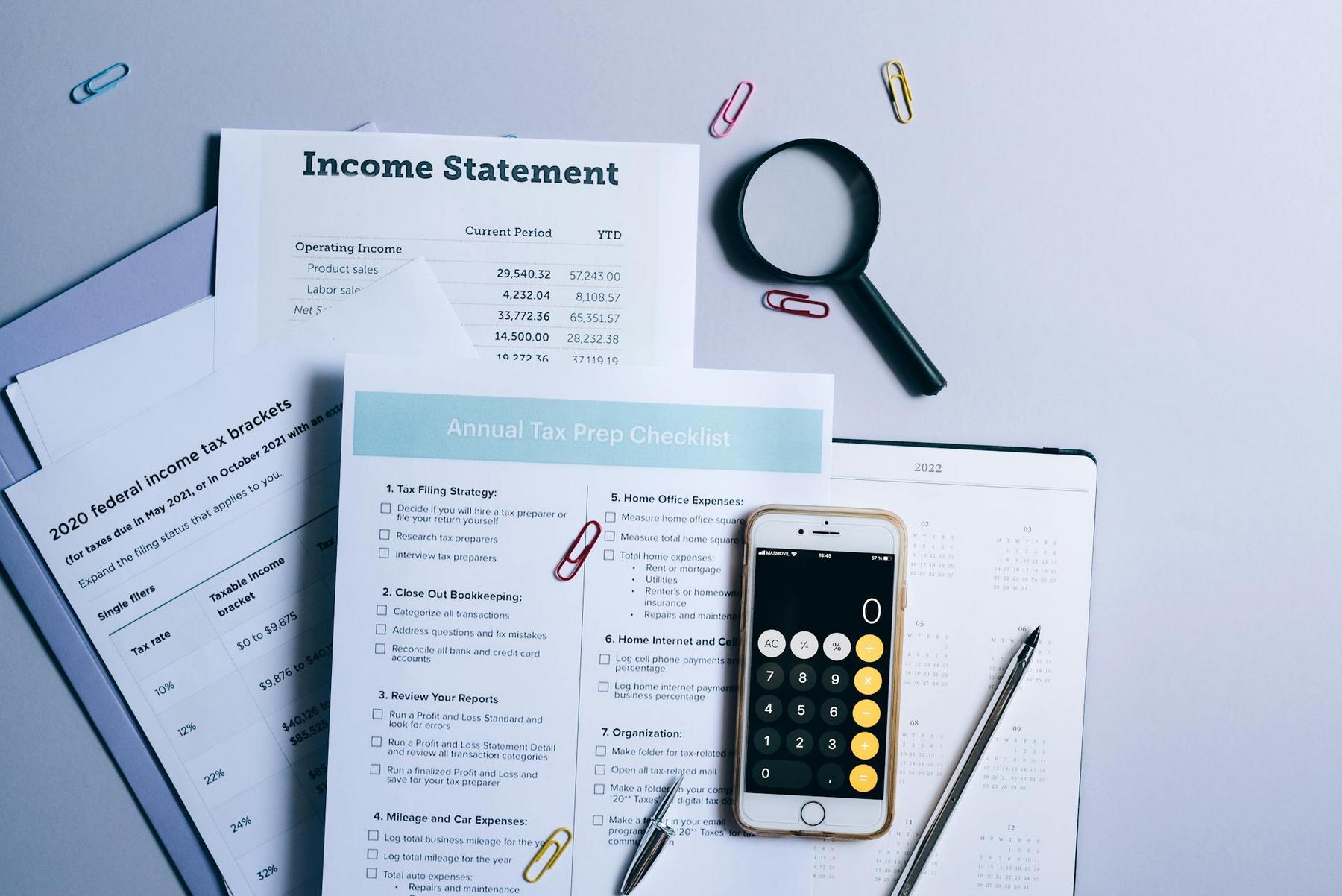 A desk with financial documents, a tax prep checklist, an income statement, a smartphone calculator, a magnifying glass, paper clips, a pen, and a planner.