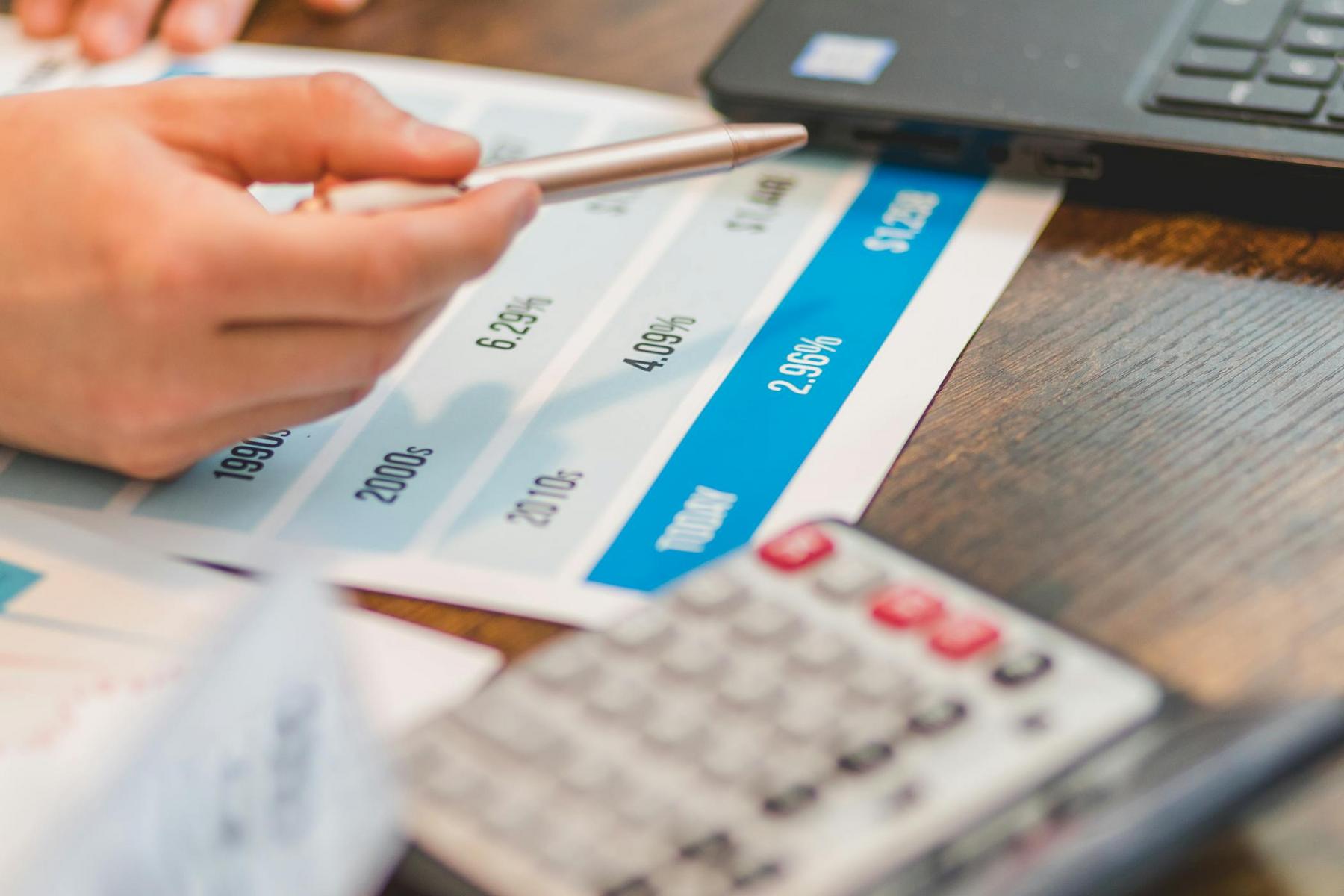 A hand holding a pen points at a printed chart with percentages, next to a calculator and a laptop on a wooden desk.