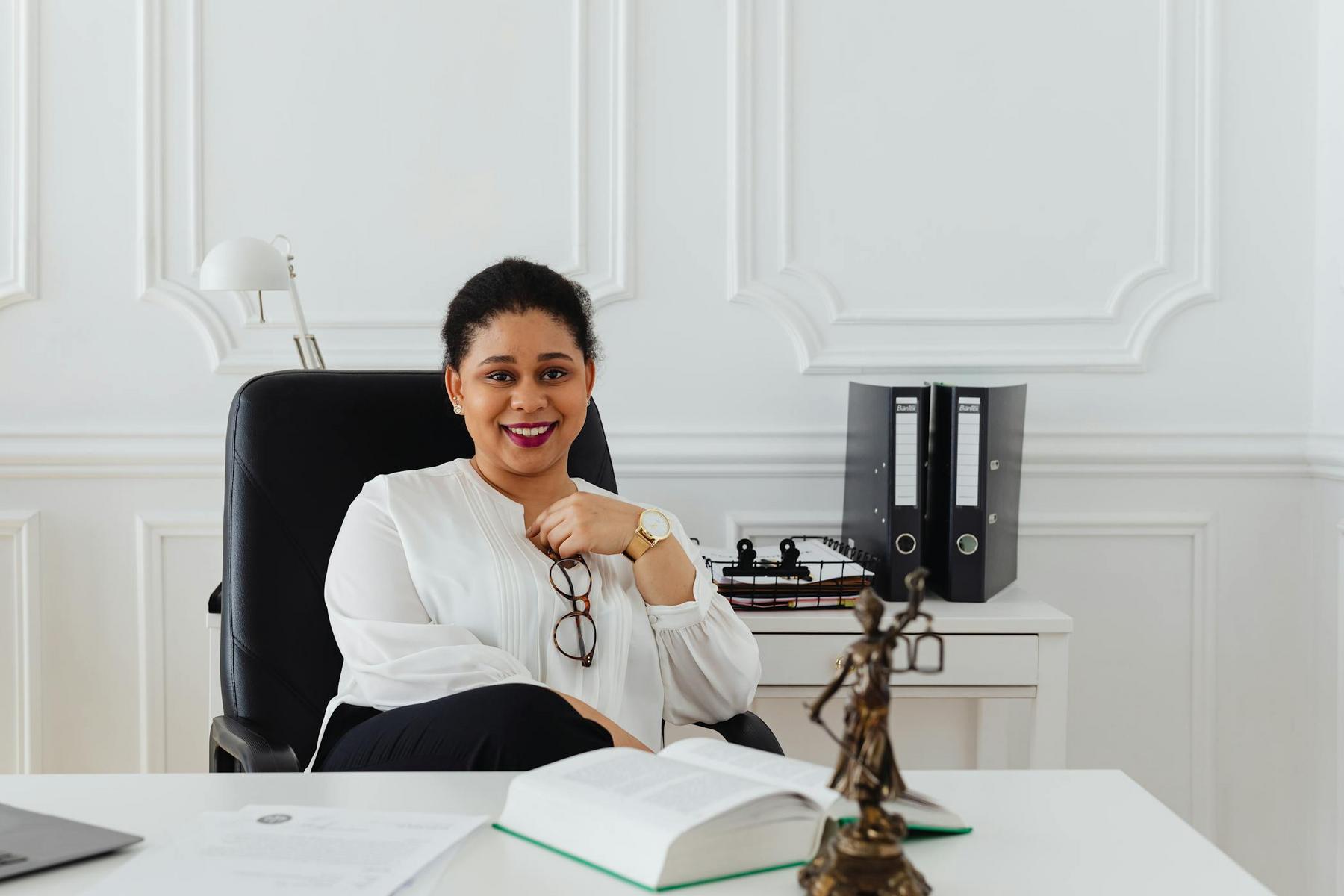 A woman sits at a desk in an office, holding eyeglasses and smiling. There is an open book, papers, and a Lady Justice statue on the desk, with binders on a shelf behind her.