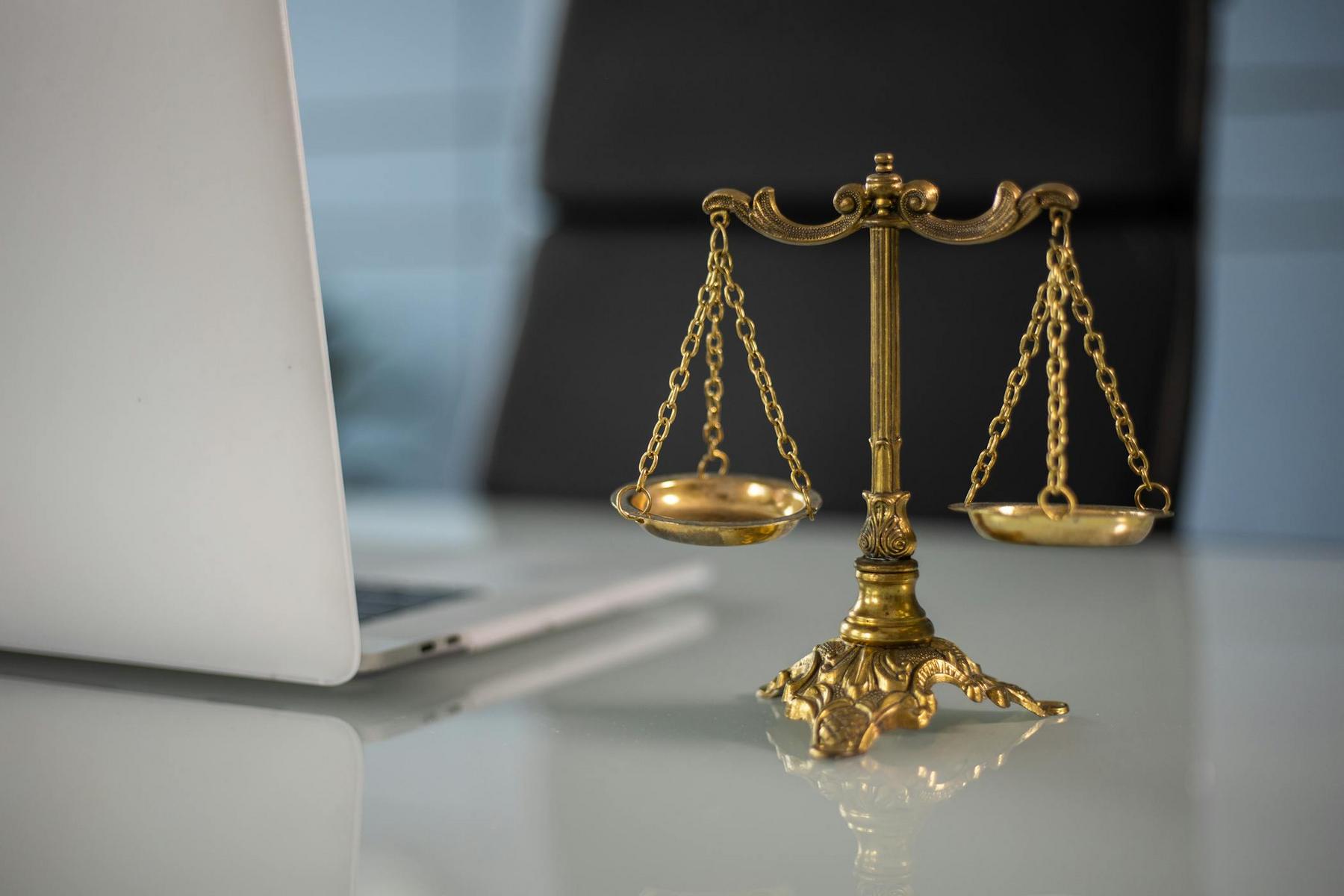 A brass balance scale sits on a glass desk next to an open laptop in an office setting.