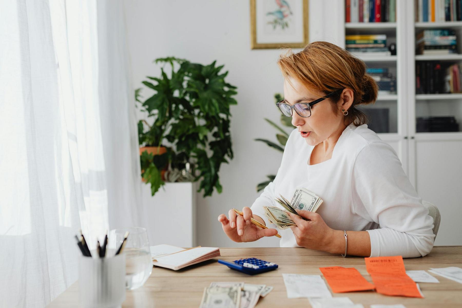 A woman in glasses sits at a table counting dollar bills with receipts, a notebook, and a calculator in front of her in a well-lit room.