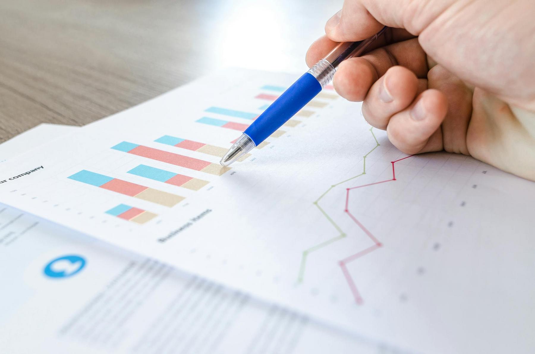 A hand holding a pen points to bar and line graphs on printed documents, indicating data analysis or review of business statistics.