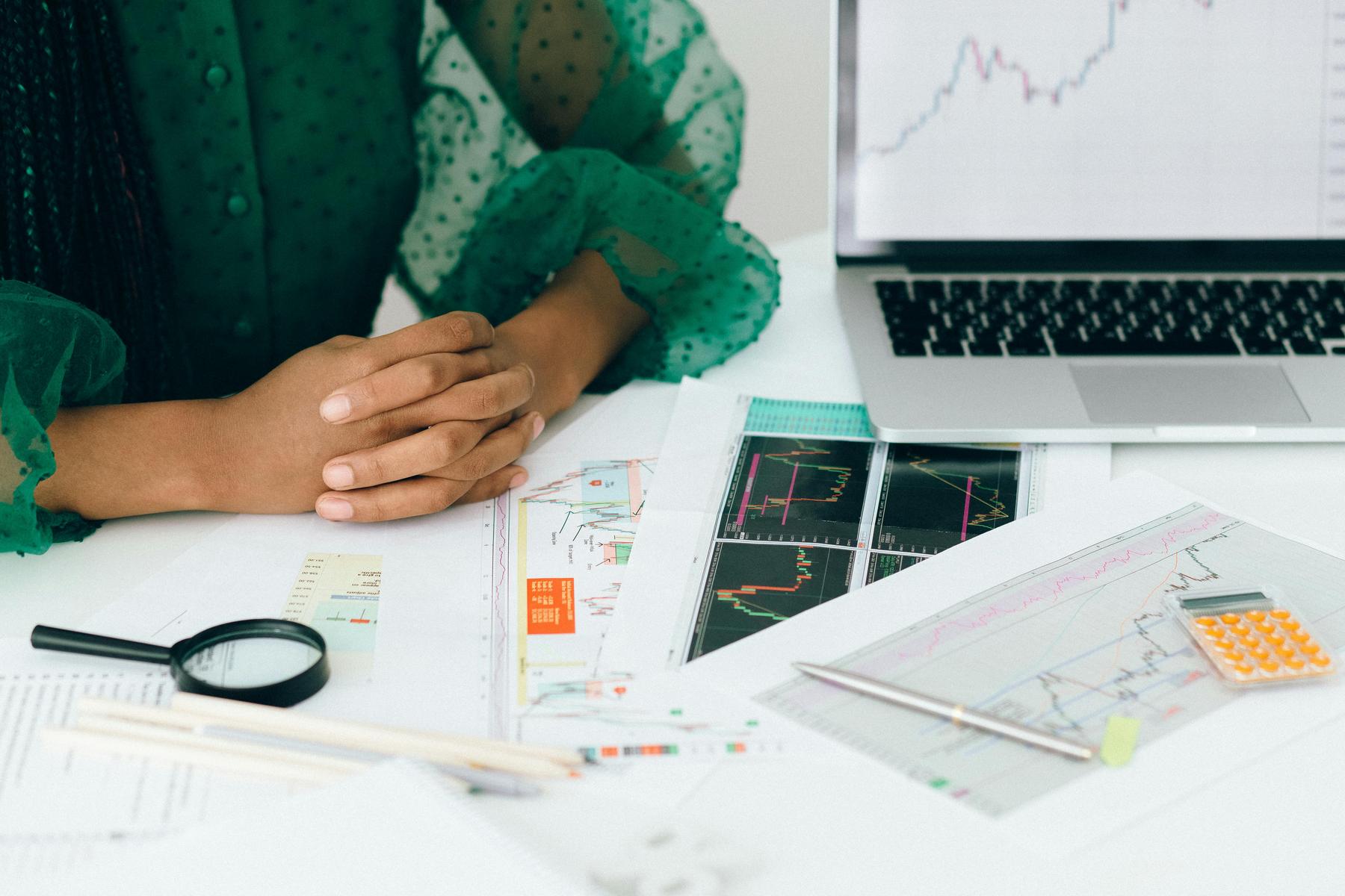 Person sitting at a desk with hands folded, surrounded by financial charts, graphs, a magnifying glass, a pen, a calculator, and a laptop displaying a rising graph.