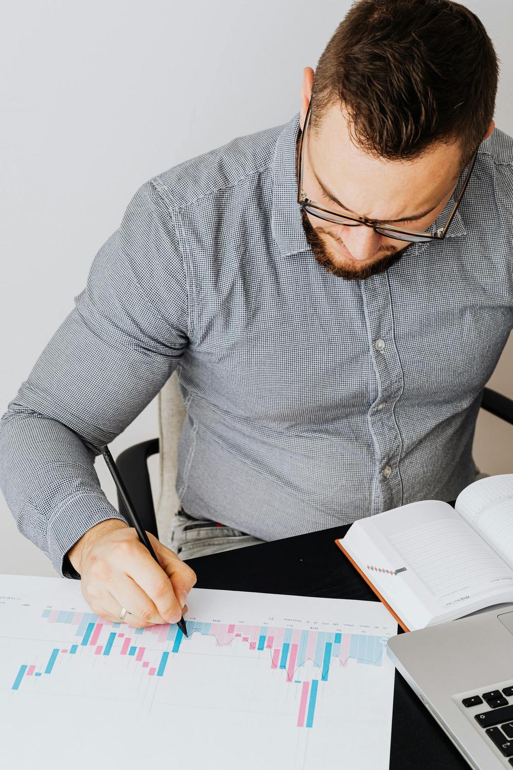A man with glasses analyzes a printed bar chart at a desk, holding a pen. An open notebook and a laptop are also on the desk.