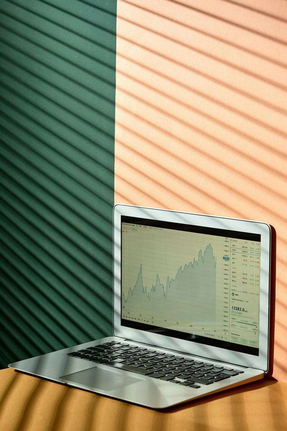 A laptop displays a line graph of financial data on its screen, with striped shadows cast on the wall and surface behind it.