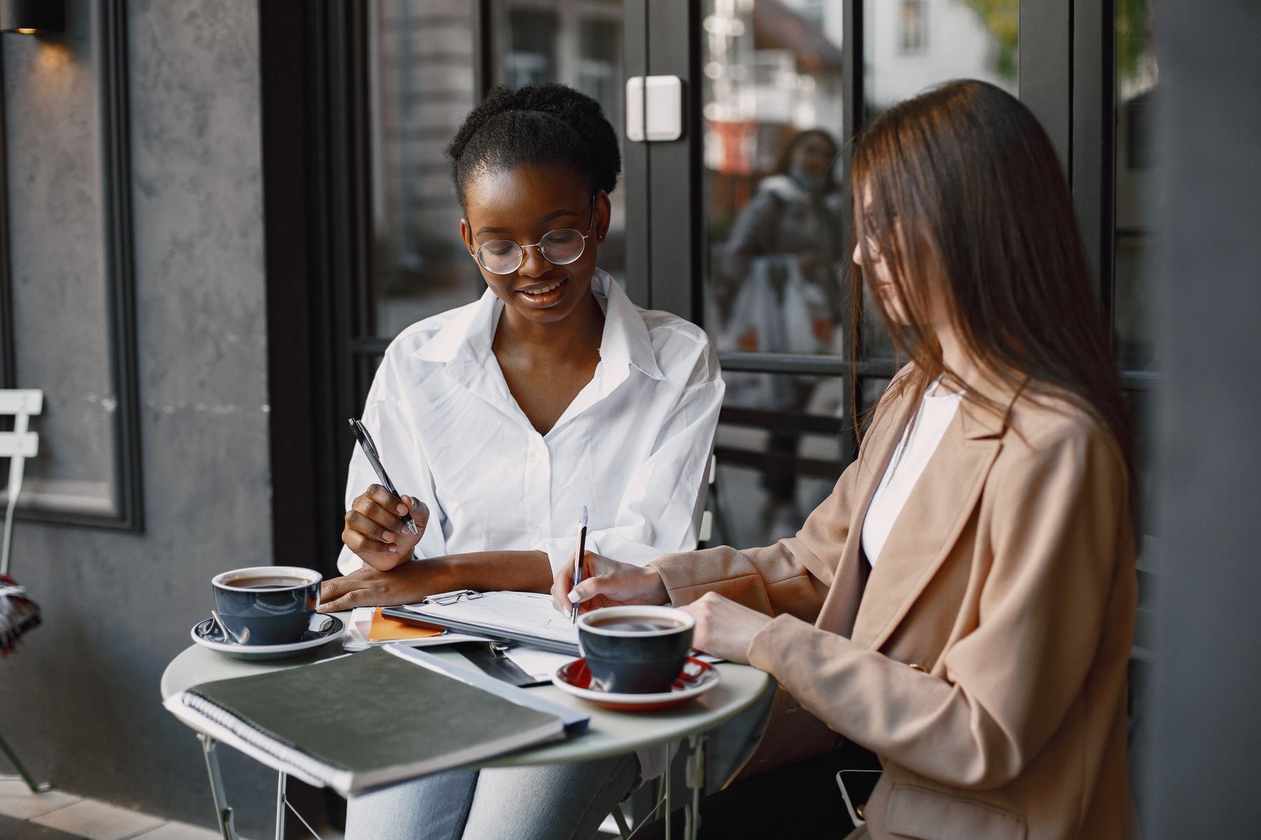 Two women at a cafe table with coffee and notebooks, discussing business at a relaxed first meeting with an accountant