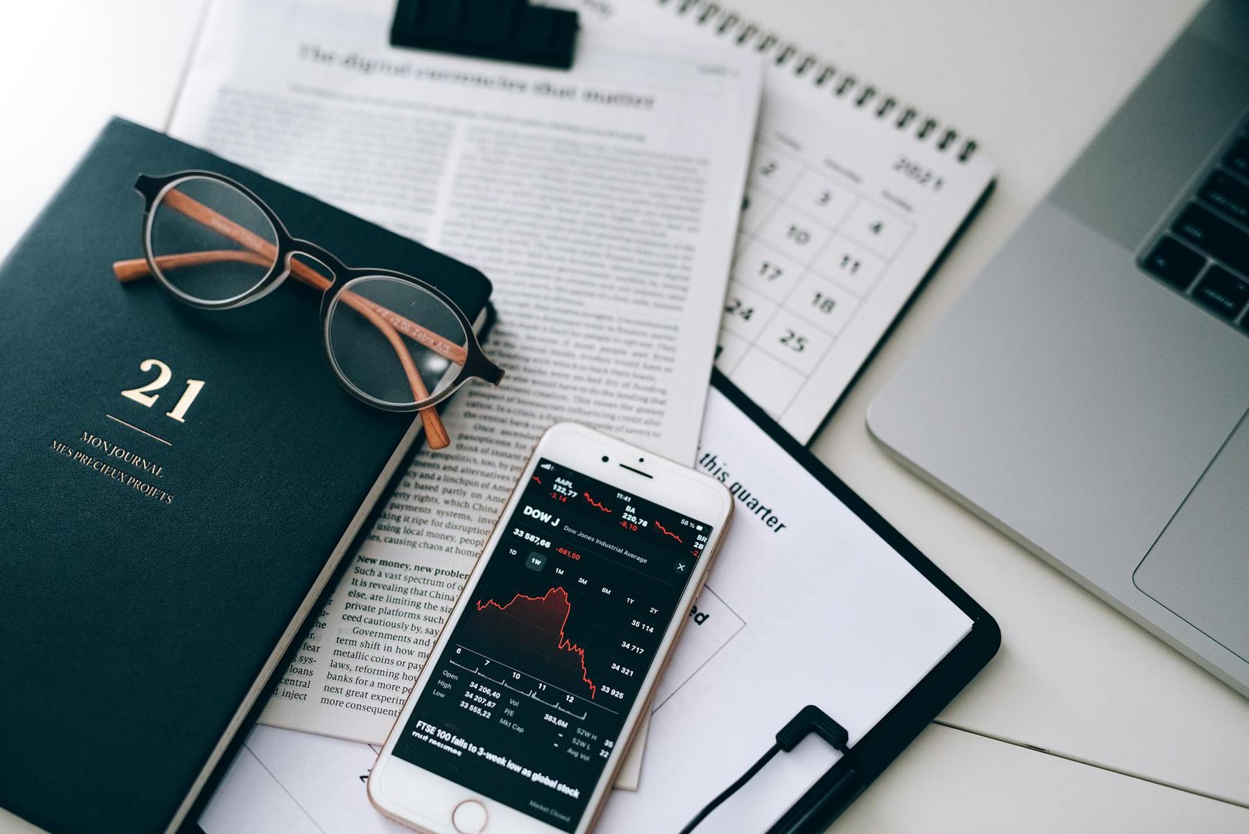 A smartphone displaying a stock chart rests on papers, a calendar, a notebook labeled "21," a pair of eyeglasses, and a laptop on a white desk.