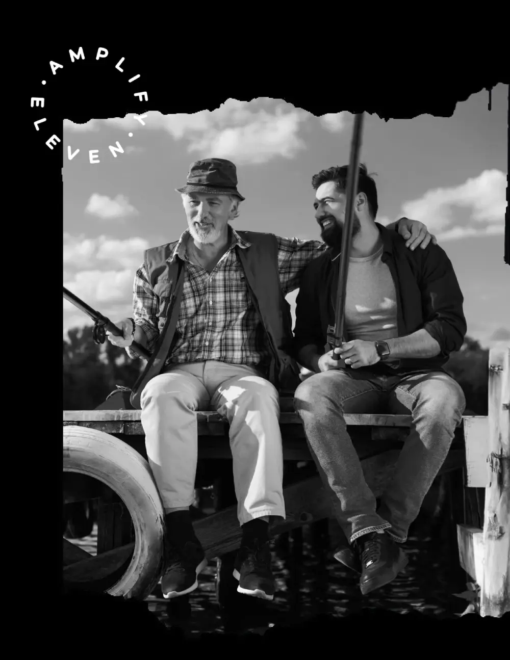 Two men sitting on a wooden dock fishing, one has his arm around the other.