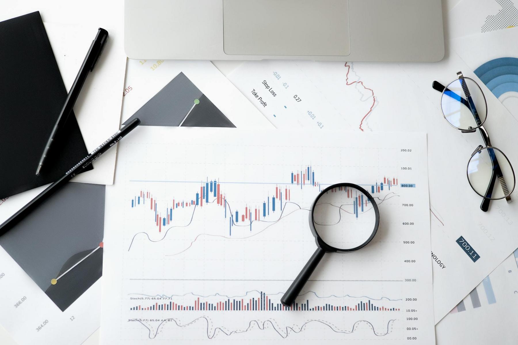 A magnifying glass rests on a printed financial chart surrounded by documents, a pen, notebook, eyeglasses, and a laptop on a white desk.