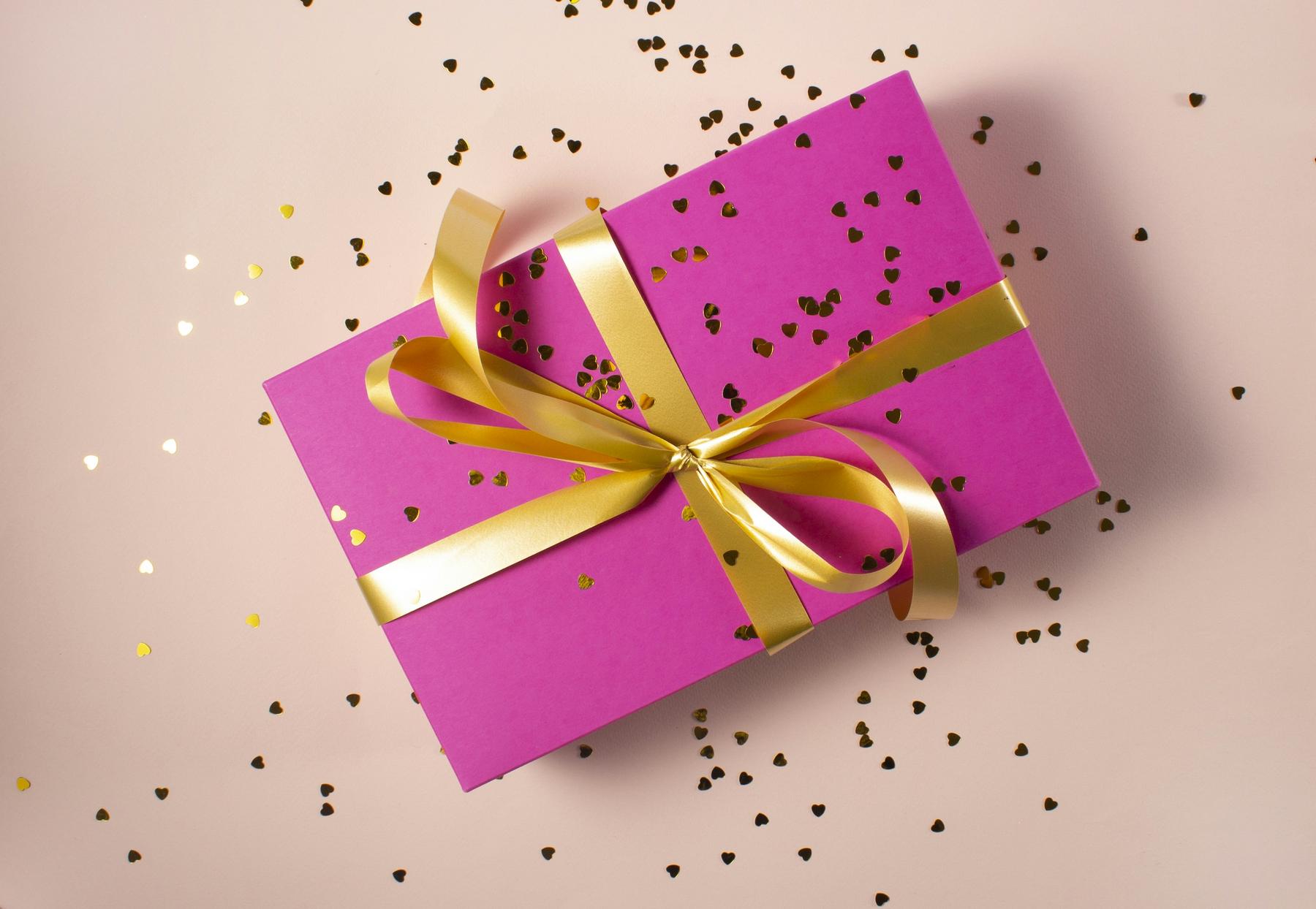 A pink gift box with a gold ribbon is surrounded by small, scattered confetti on a light-colored surface.