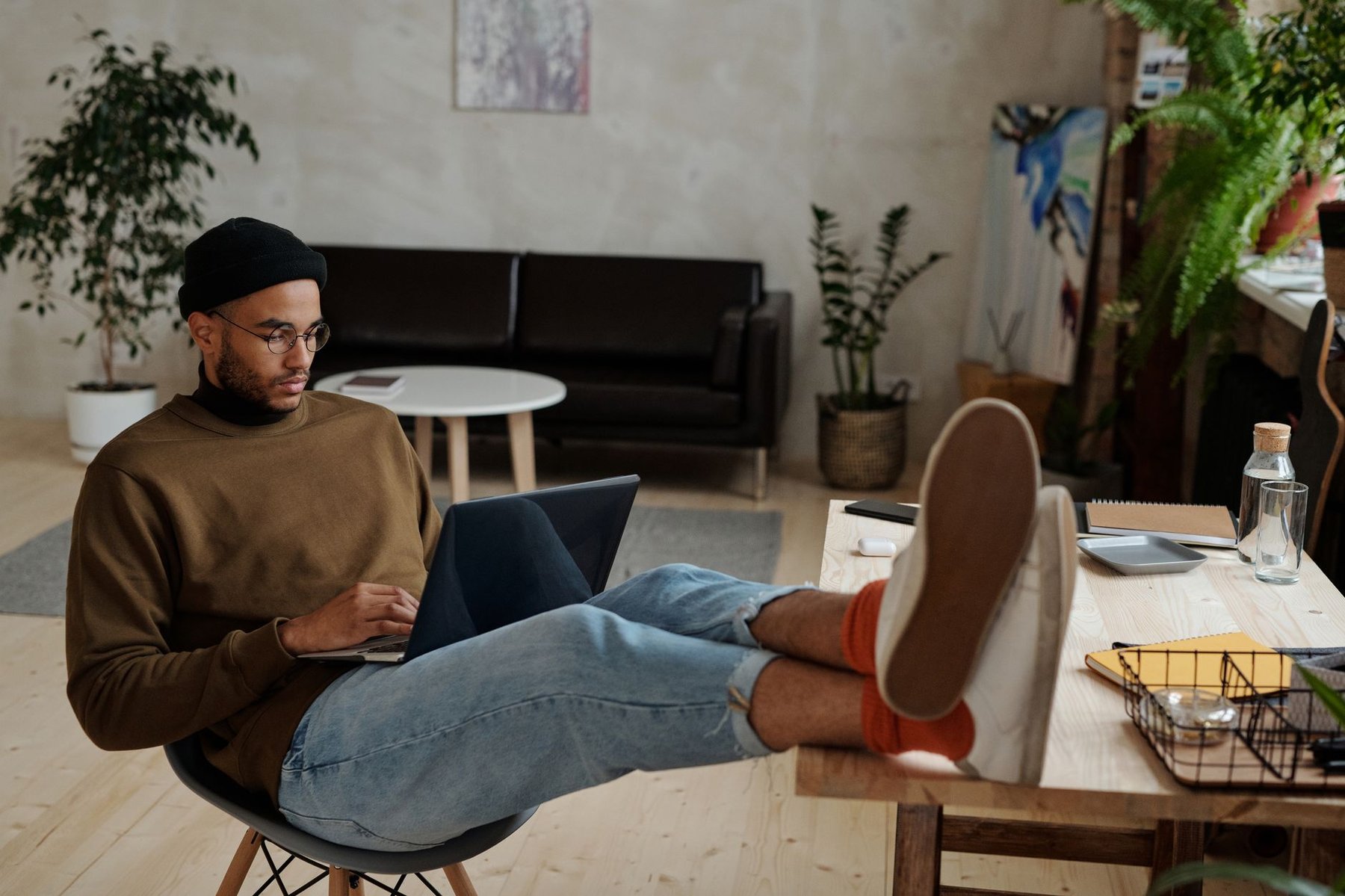 Relaxed freelancer with feet up on a wooden desk working on a laptop in a bright, plant-filled creative home studio
