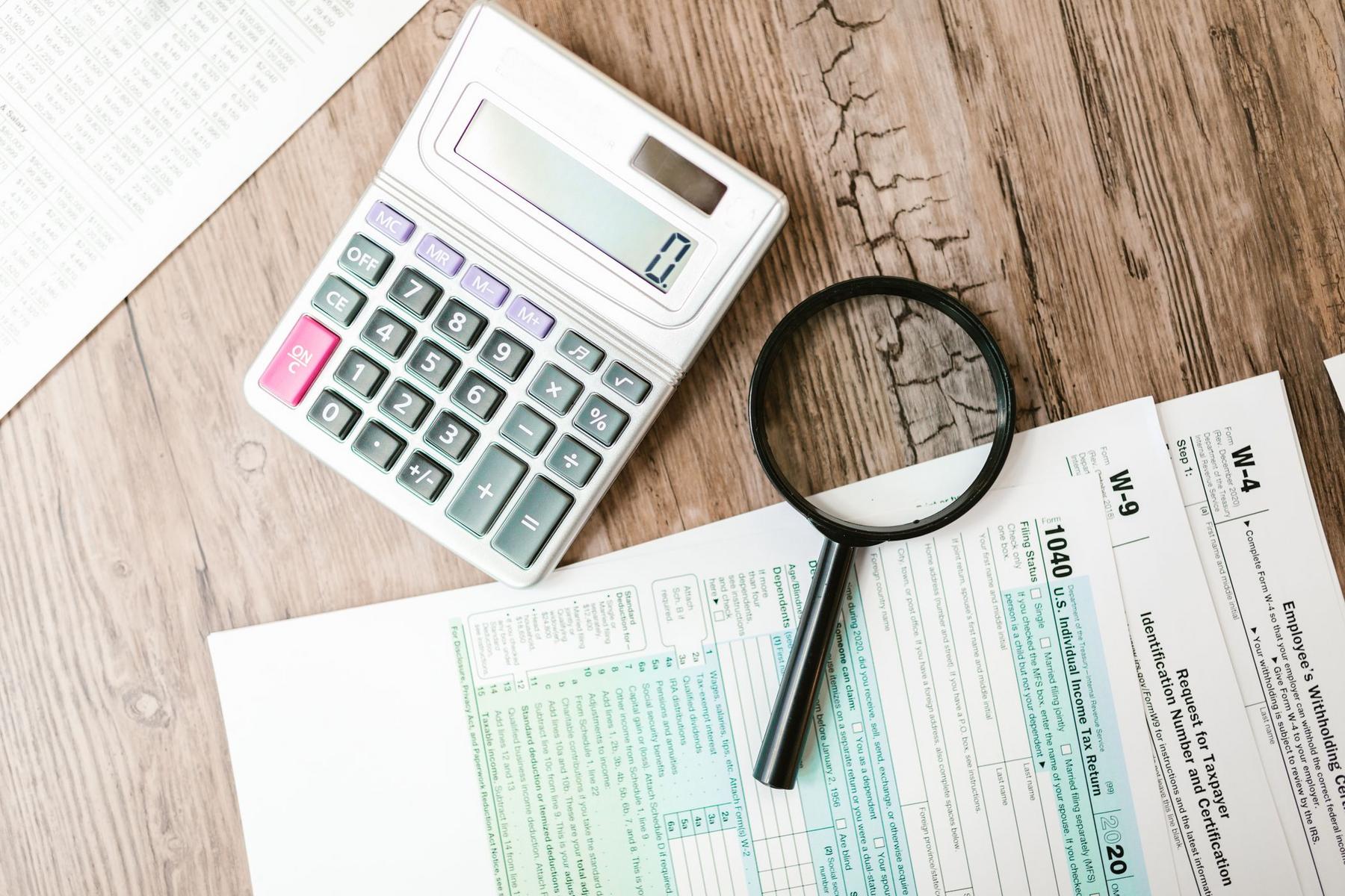 A calculator, magnifying glass, and various tax forms are arranged on a wooden surface.