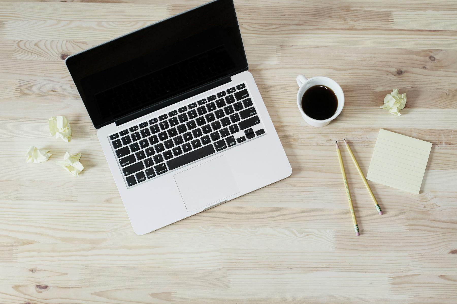 A laptop on a wooden desk with a cup of coffee, two pencils, yellow sticky notes, and crumpled paper balls.