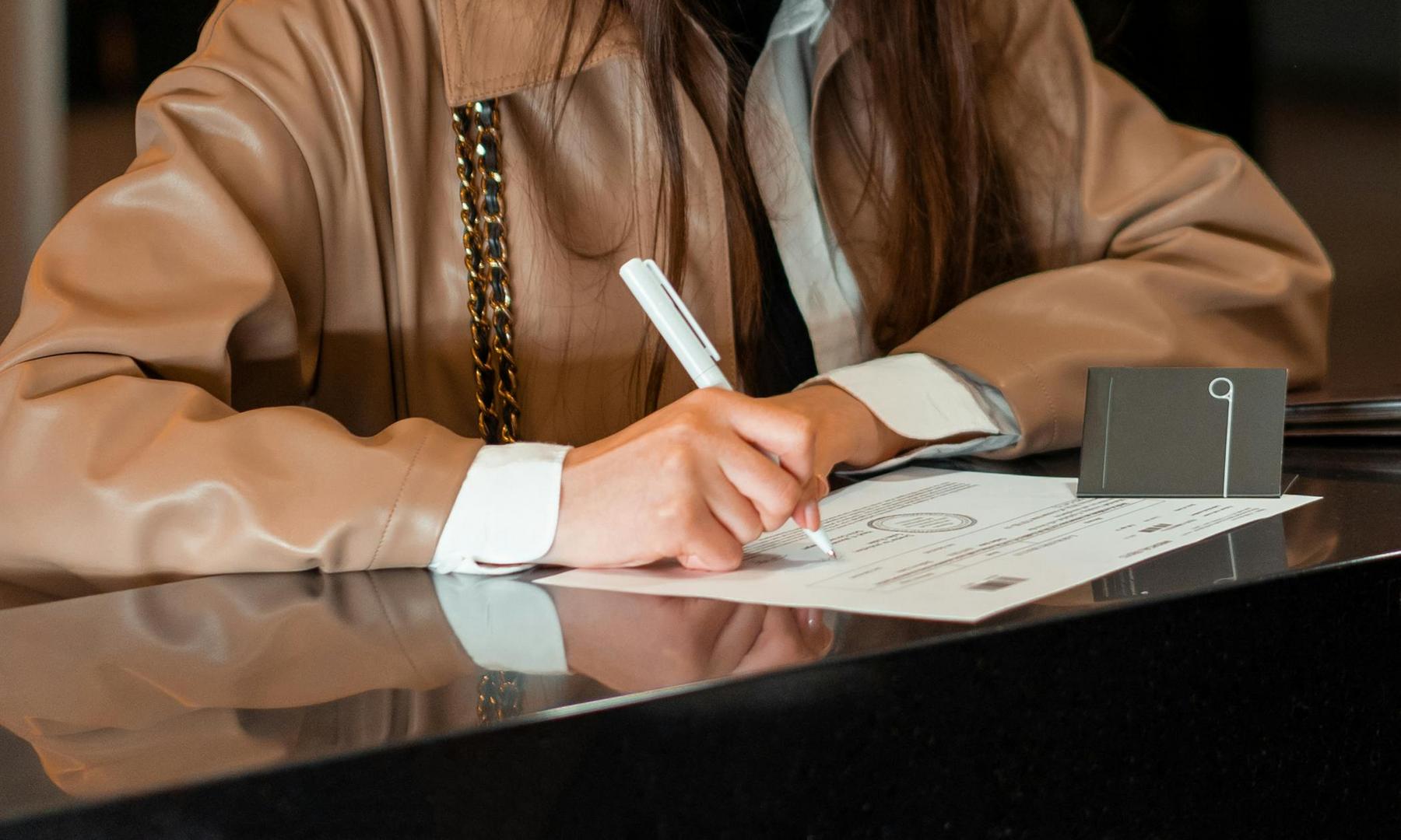A person wearing a tan coat fills out a form with a pen on a counter; a keycard holder is placed beside the form.