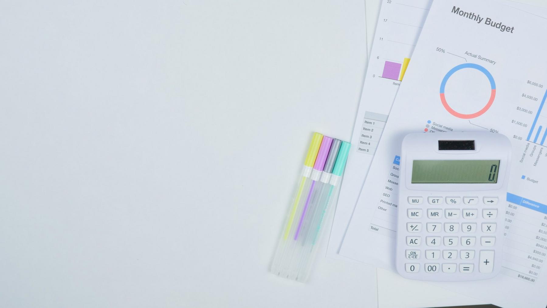 White calculator, colorful pens, and monthly budget reports with charts and graphs on a white desk, with ample empty space on the left side.