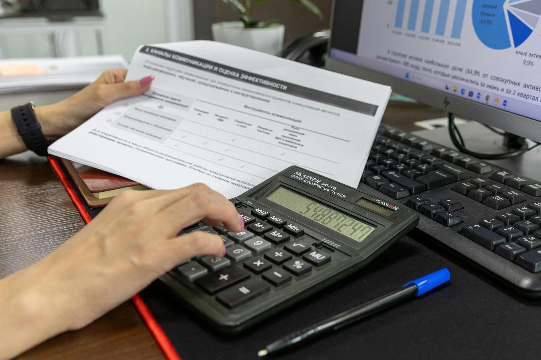 Person holding a document and using a calculator next to a computer keyboard and a monitor displaying graphs.
