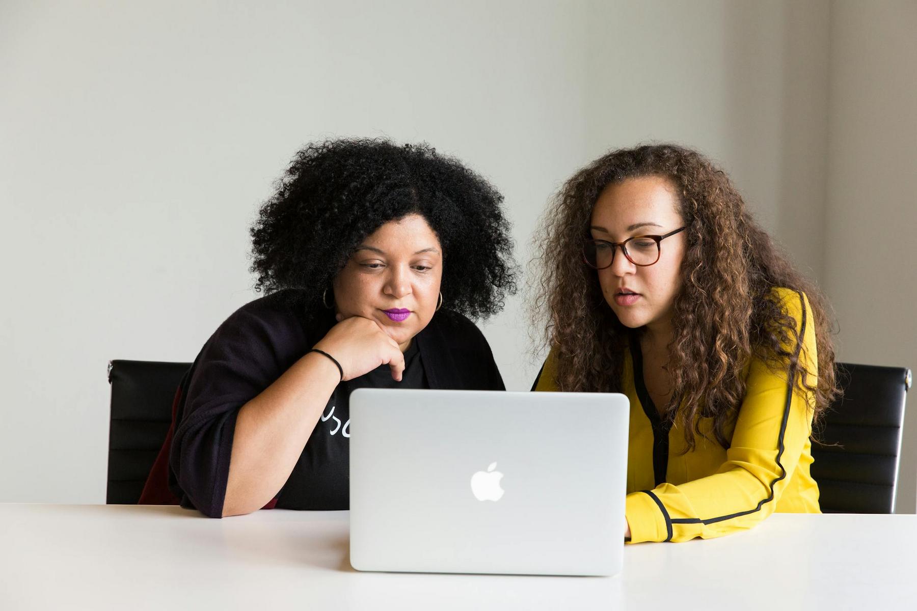 Two women sitting at a table, looking at a laptop screen together in a bright, minimalist office setting.