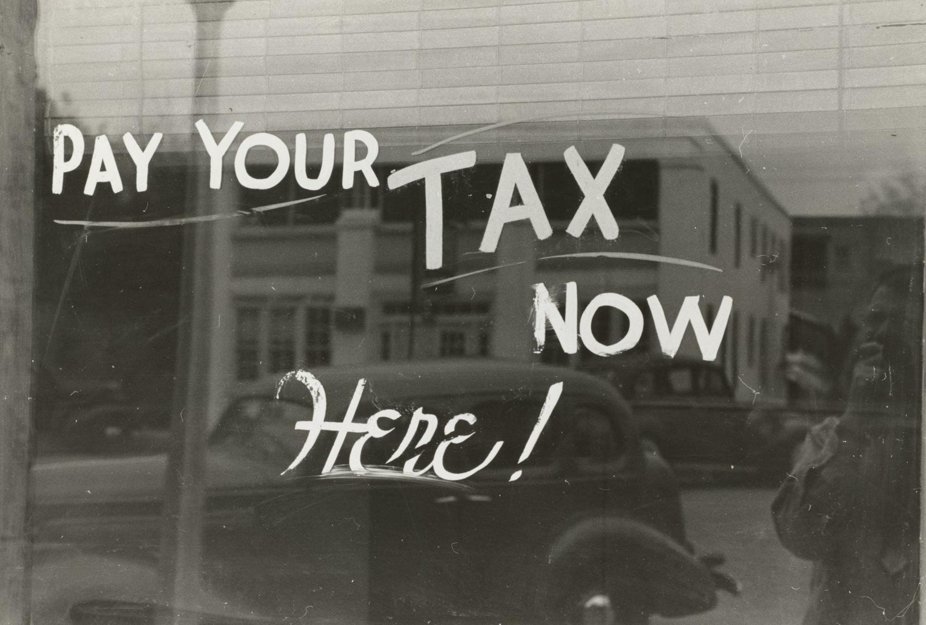 A window sign reads "Pay Your Tax Now Here!" with a reflection of a vintage car and building in the background.