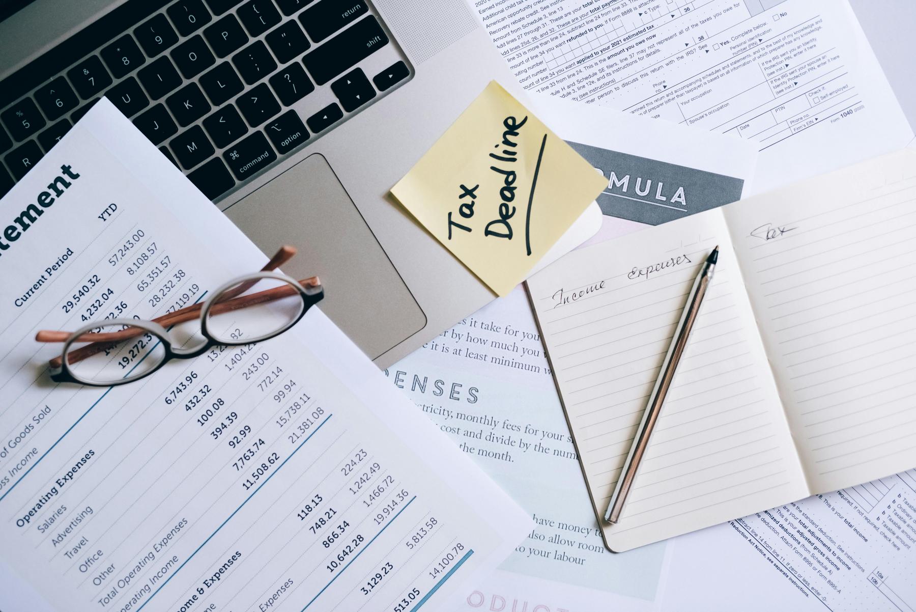 A laptop, financial documents, eyeglasses, a notebook labeled “Income/Expenses,” pen, and a sticky note reading “Tax Deadline” are arranged on a desk.