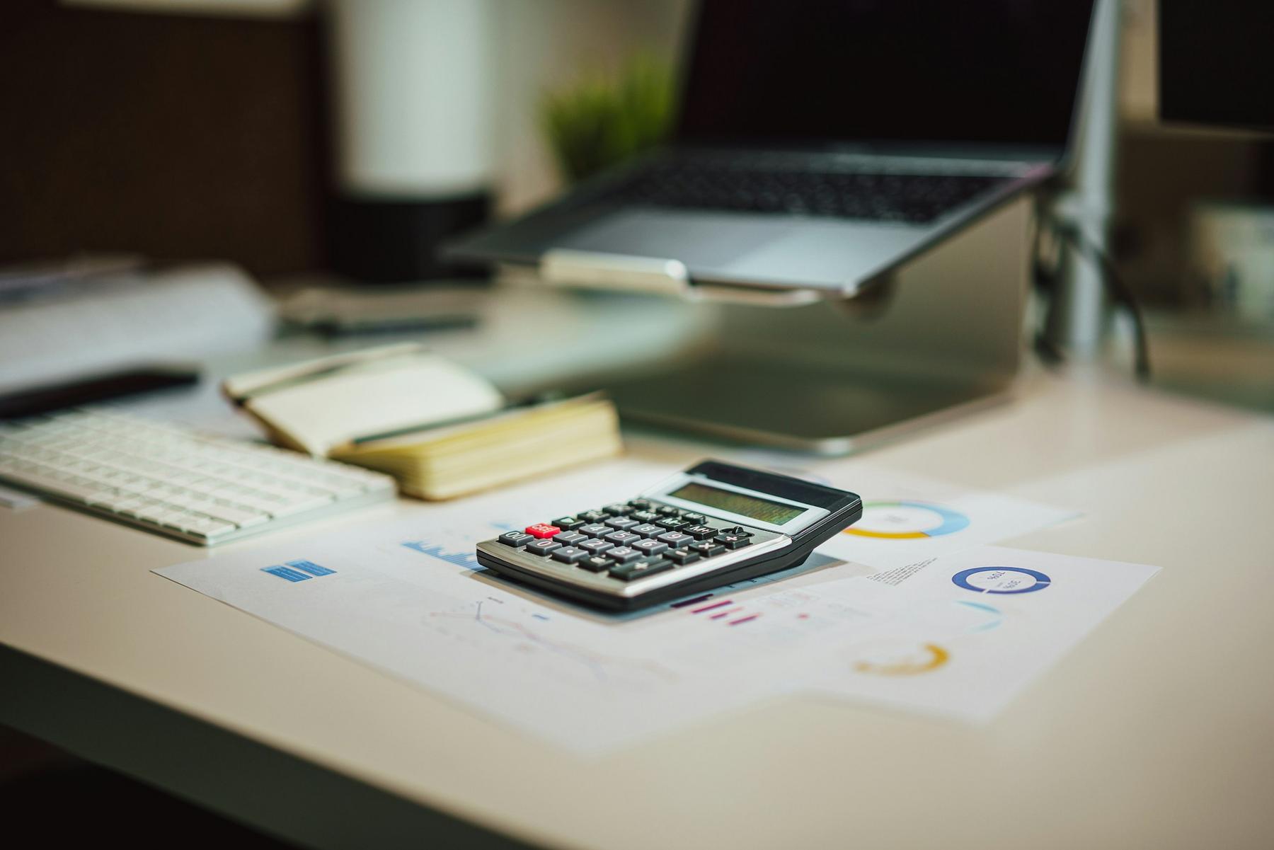 A calculator sits on top of charts and graphs on a desk with a keyboard, open notebook, and a laptop on a stand in the background.