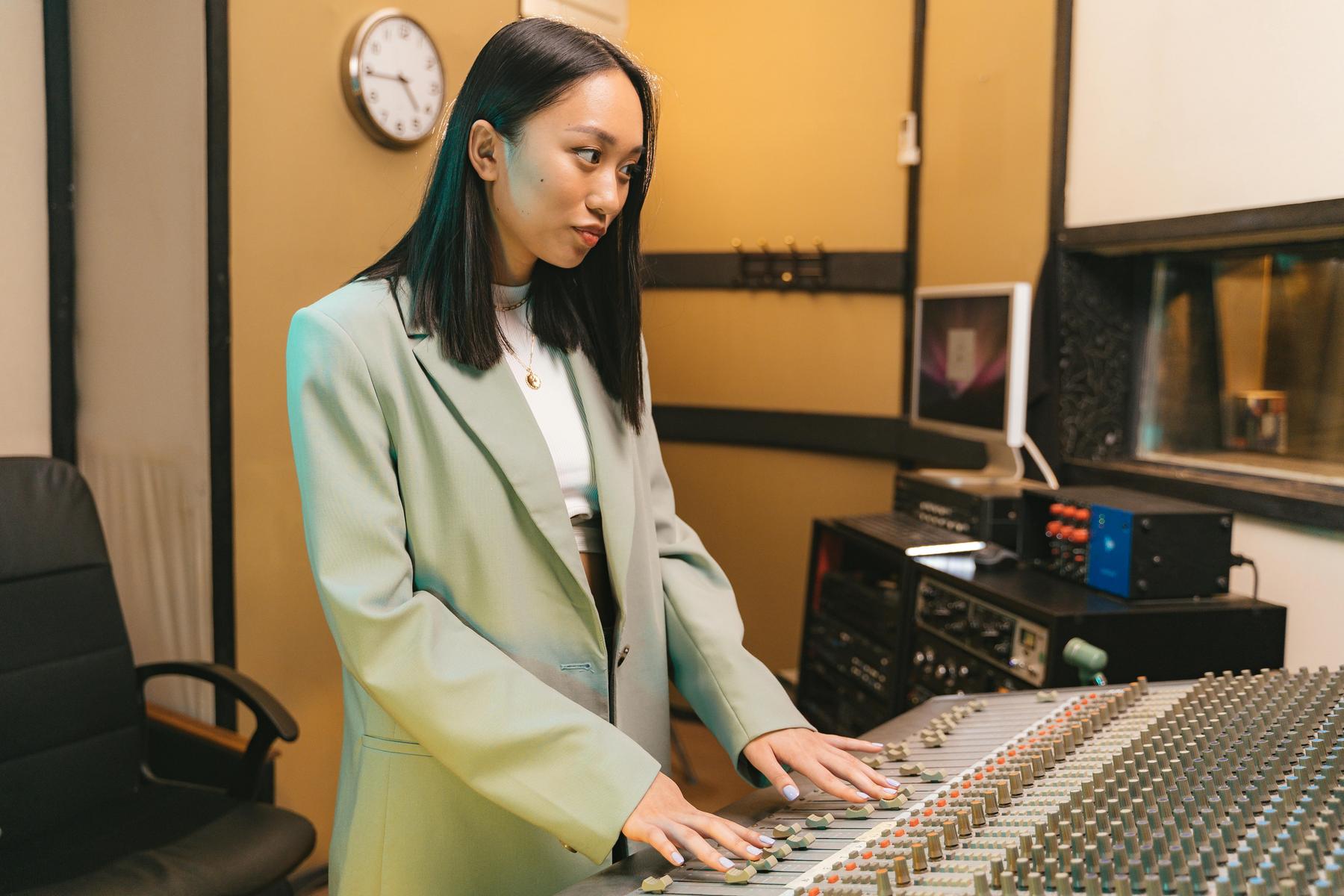A woman in a green blazer stands at an audio mixing console in a recording studio, adjusting the controls.