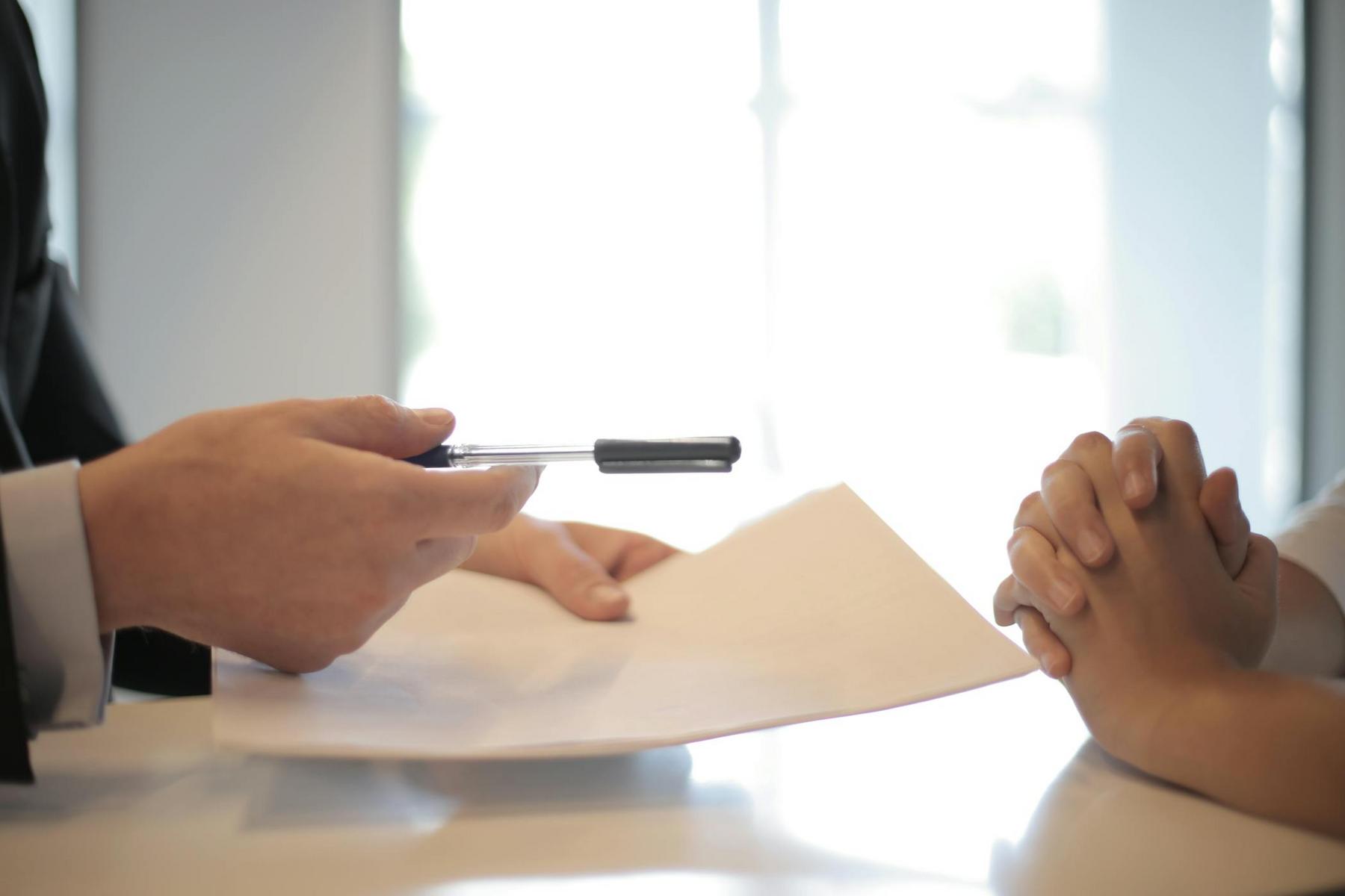 A person hands a pen and document to another person with clasped hands, suggesting a signing or agreement process at a desk.
