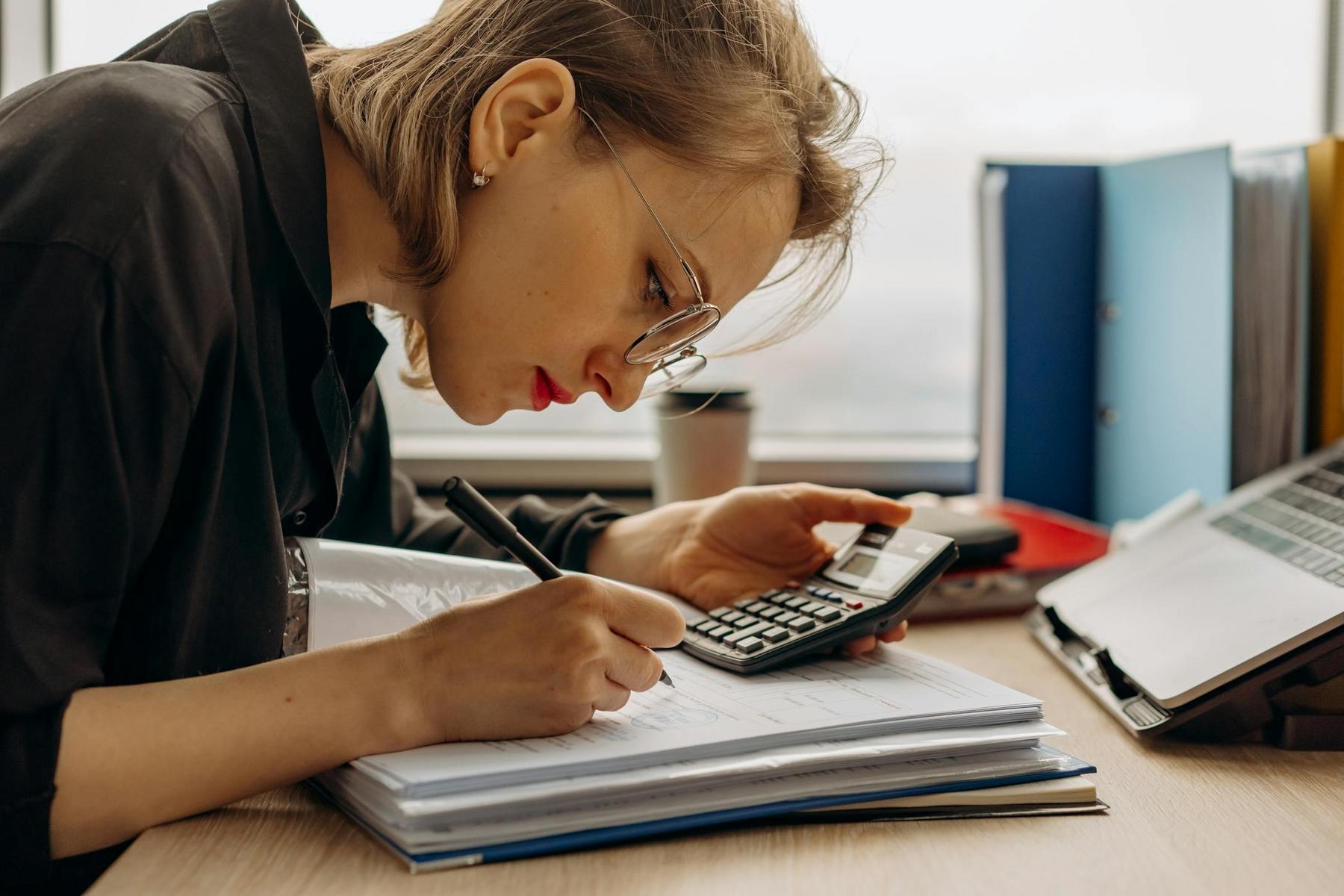 Person wearing glasses calculates figures with a calculator and pen, writing in a notebook at a desk with folders, a tablet, and a cup.