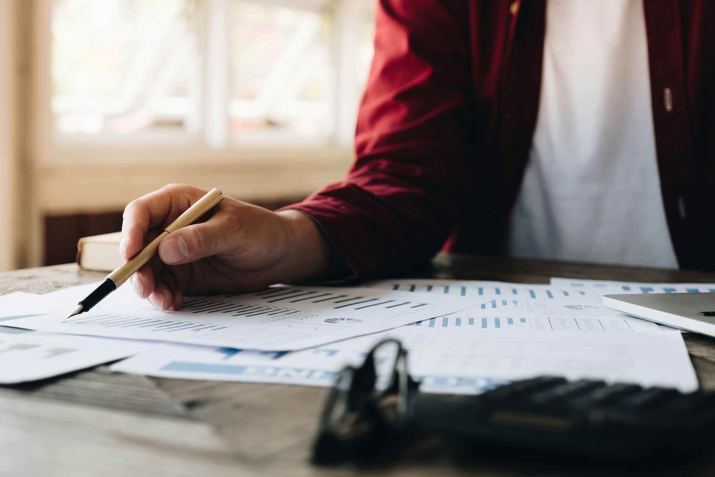 Person writing on documents at a desk, pen in hand, surrounded by glasses and a calculator. In the well-lit room, they ponder how to find your tax file number efficiently and accurately.