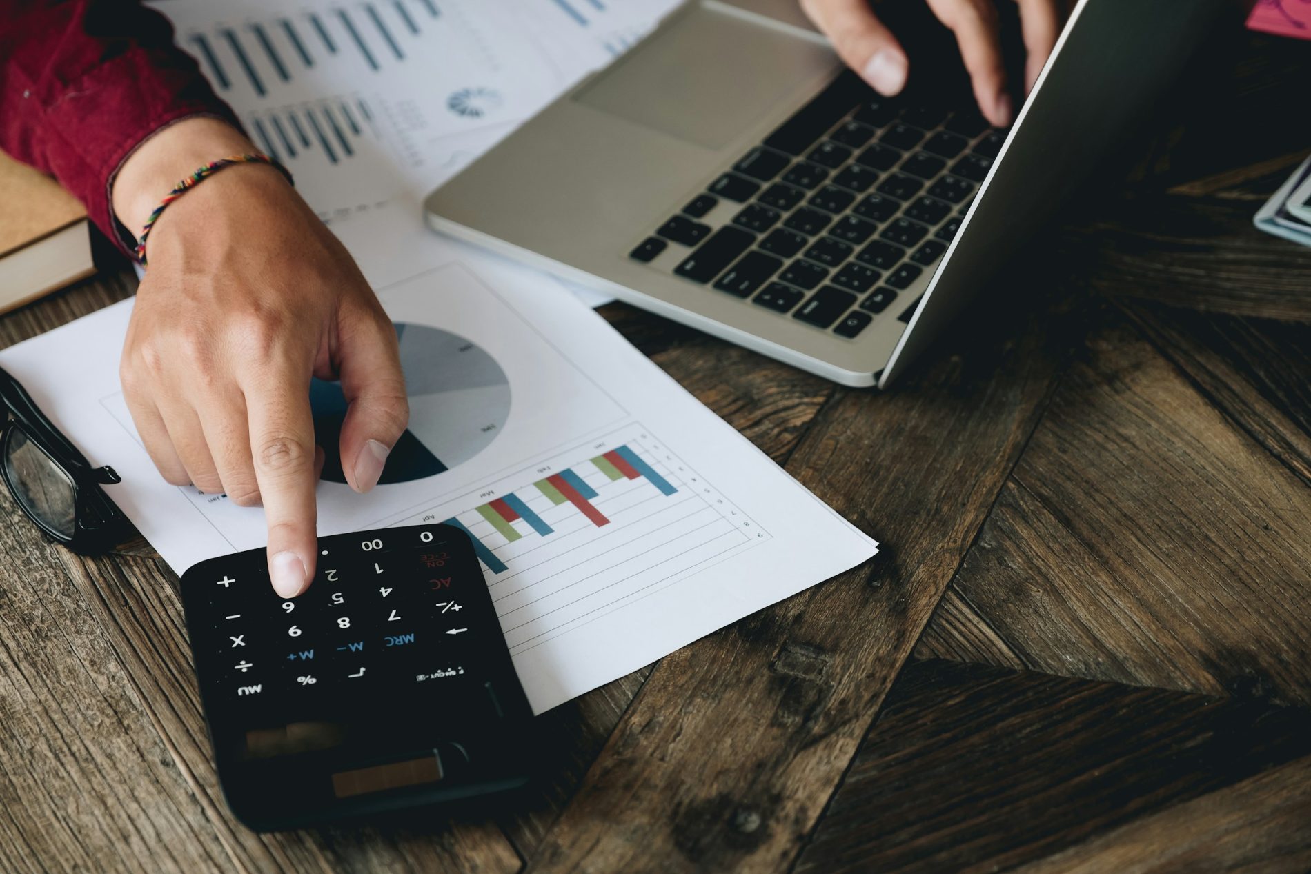 Amidst printed charts and graphs on a wooden table, a person uses a calculator and laptop to navigate financial duties, perhaps even figuring out how to find your tax file number.