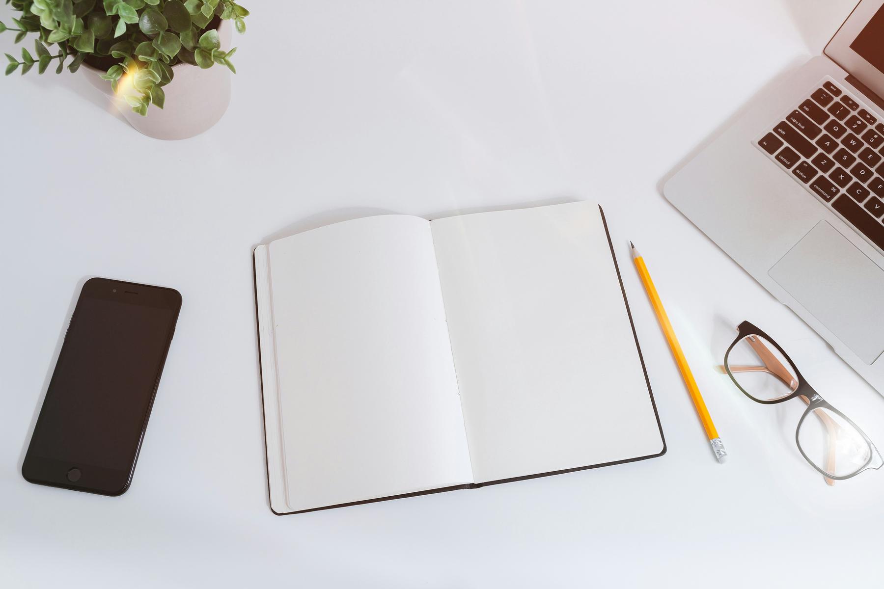 An open notebook, pencil, glasses, smartphone, and laptop are arranged on a white desk with a small potted plant.