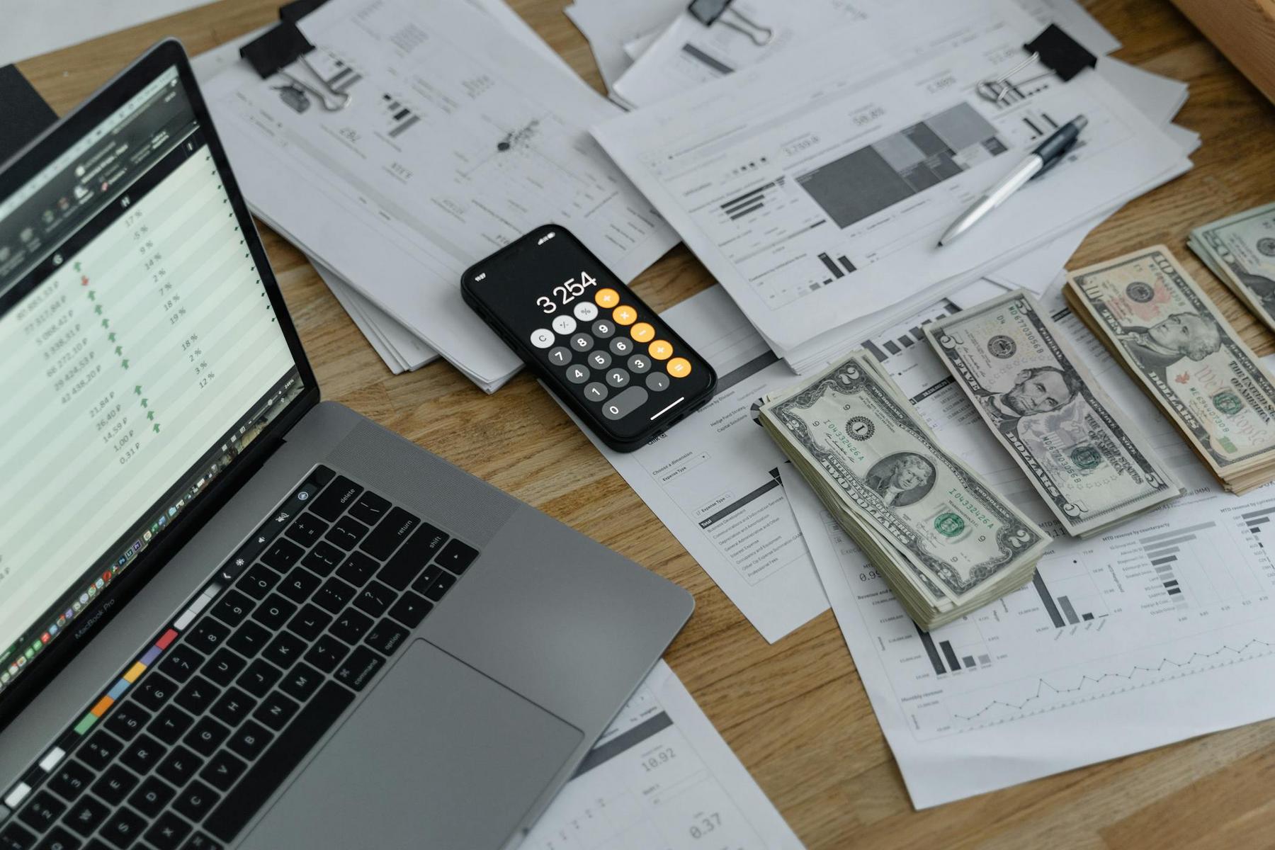 A laptop, smartphone with a calculator app, US dollar bills, and printed charts and graphs are spread out on a wooden desk.
