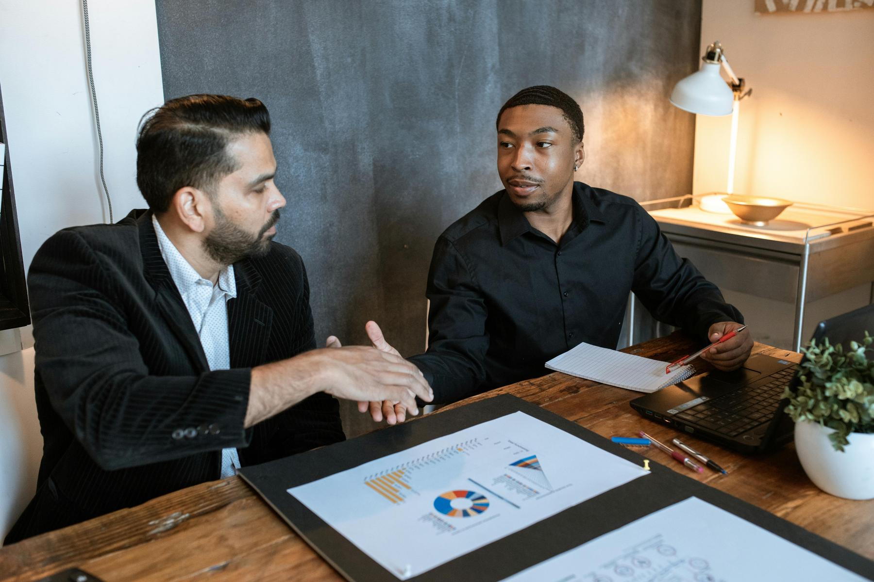 Two men sitting at a desk shake hands, with charts and a laptop in front of them, suggesting a business meeting or agreement.