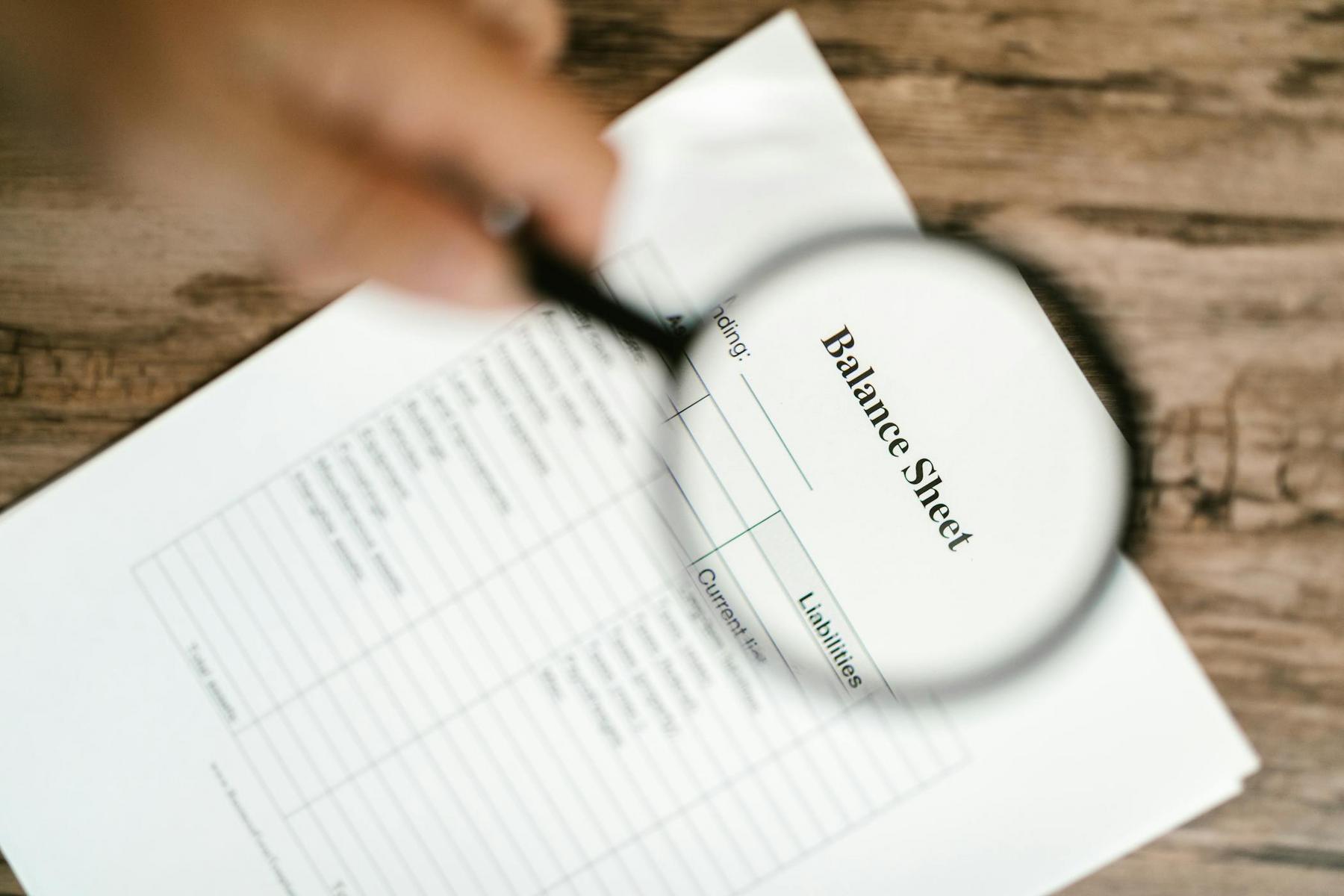 A hand holds a magnifying glass over a printed balance sheet on a wooden surface.