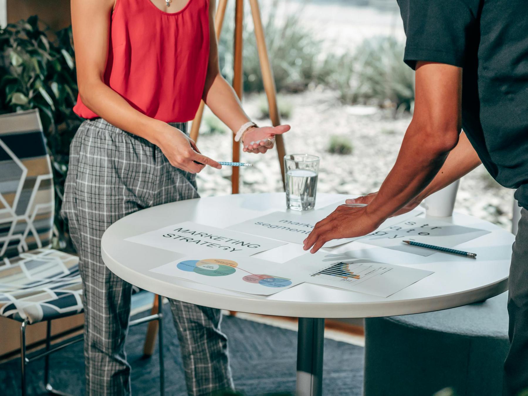 Two people stand by a round table discussing documents and charts labeled “Marketing Strategy”; a glass of water and pen are also on the table.