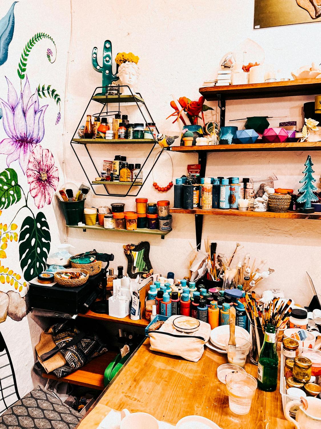 A cluttered art studio with shelves of paint jars, brushes, and supplies, a wooden table, and a colorful mural featuring flowers and leaves on the wall.
