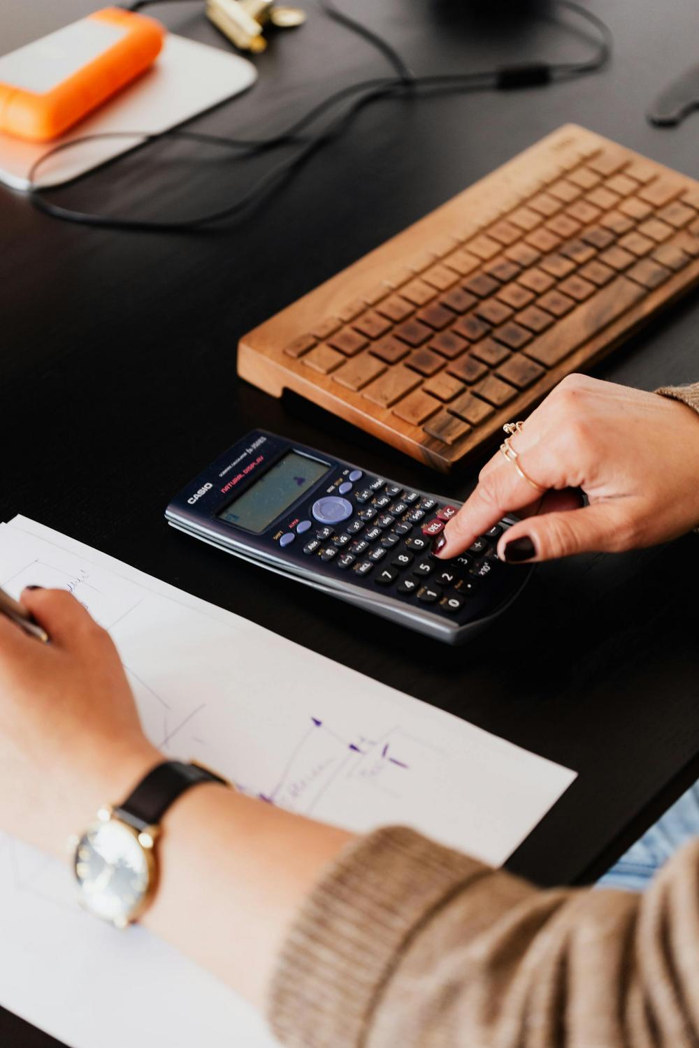 A person uses a scientific calculator beside a wooden keyboard and a sheet of paper with handwritten diagrams on a desk.