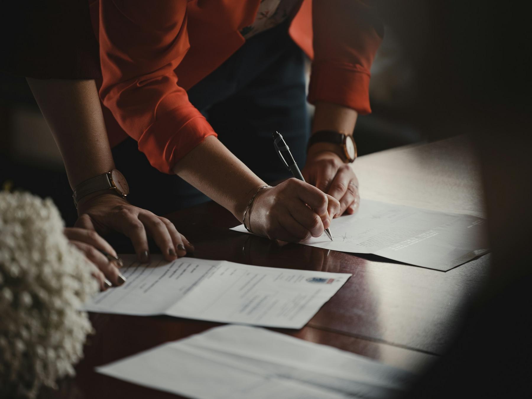 Person in a red jacket signing a document on a wooden table, with another individual's hand resting nearby and papers scattered around.