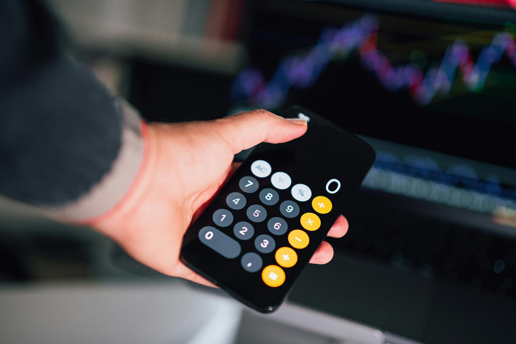 A person holding a smartphone displaying a calculator app, with a blurred trading chart visible in the background.