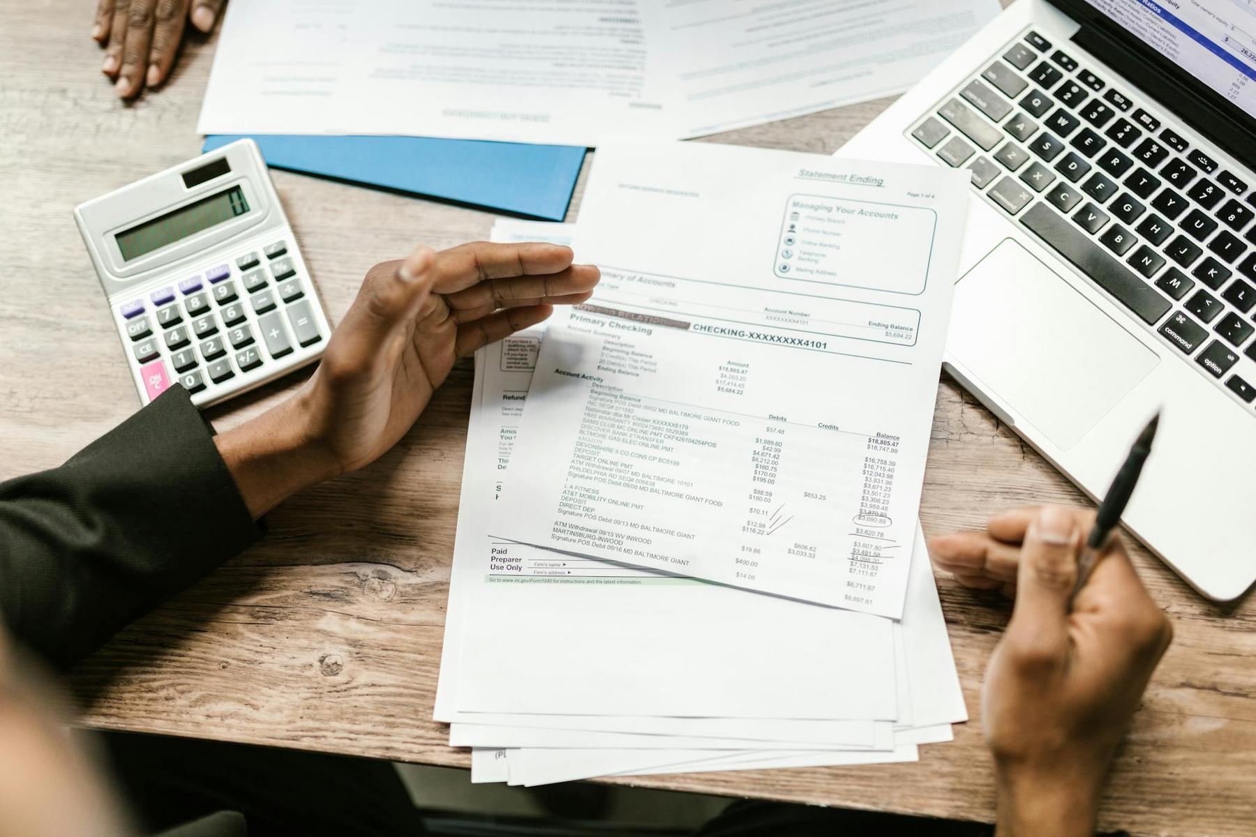 Person holding documents with financial statements, calculator, and pen on a wooden desk next to a laptop and additional paperwork.