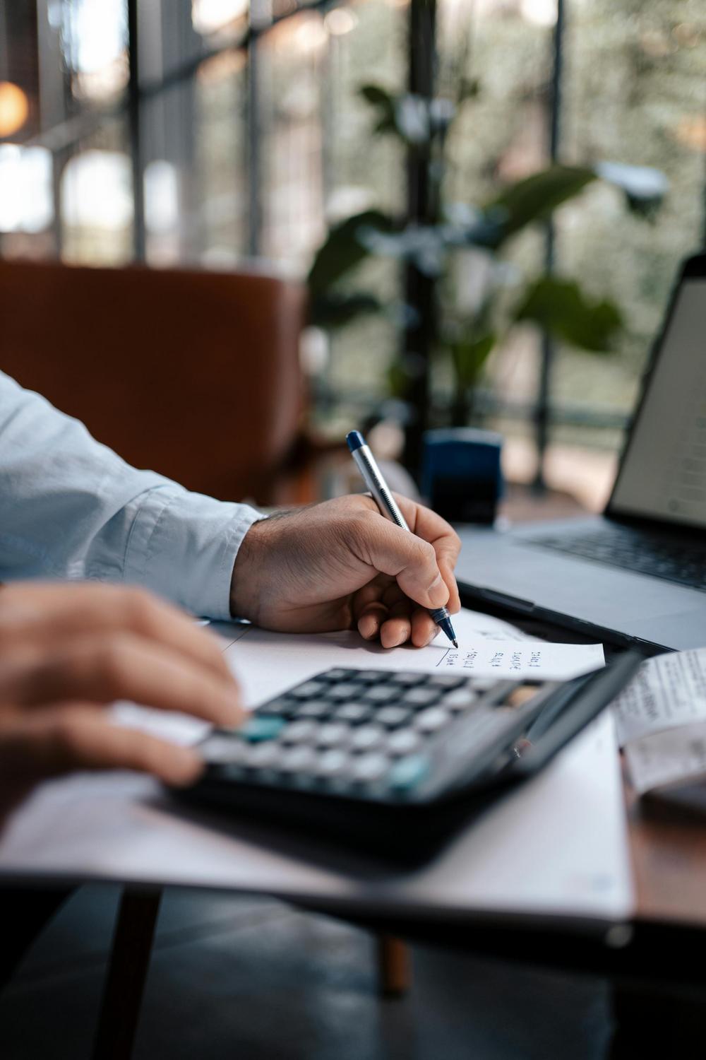 A person uses a calculator and writes on a paper at a desk with a laptop, in a modern office with large windows and indoor plants.