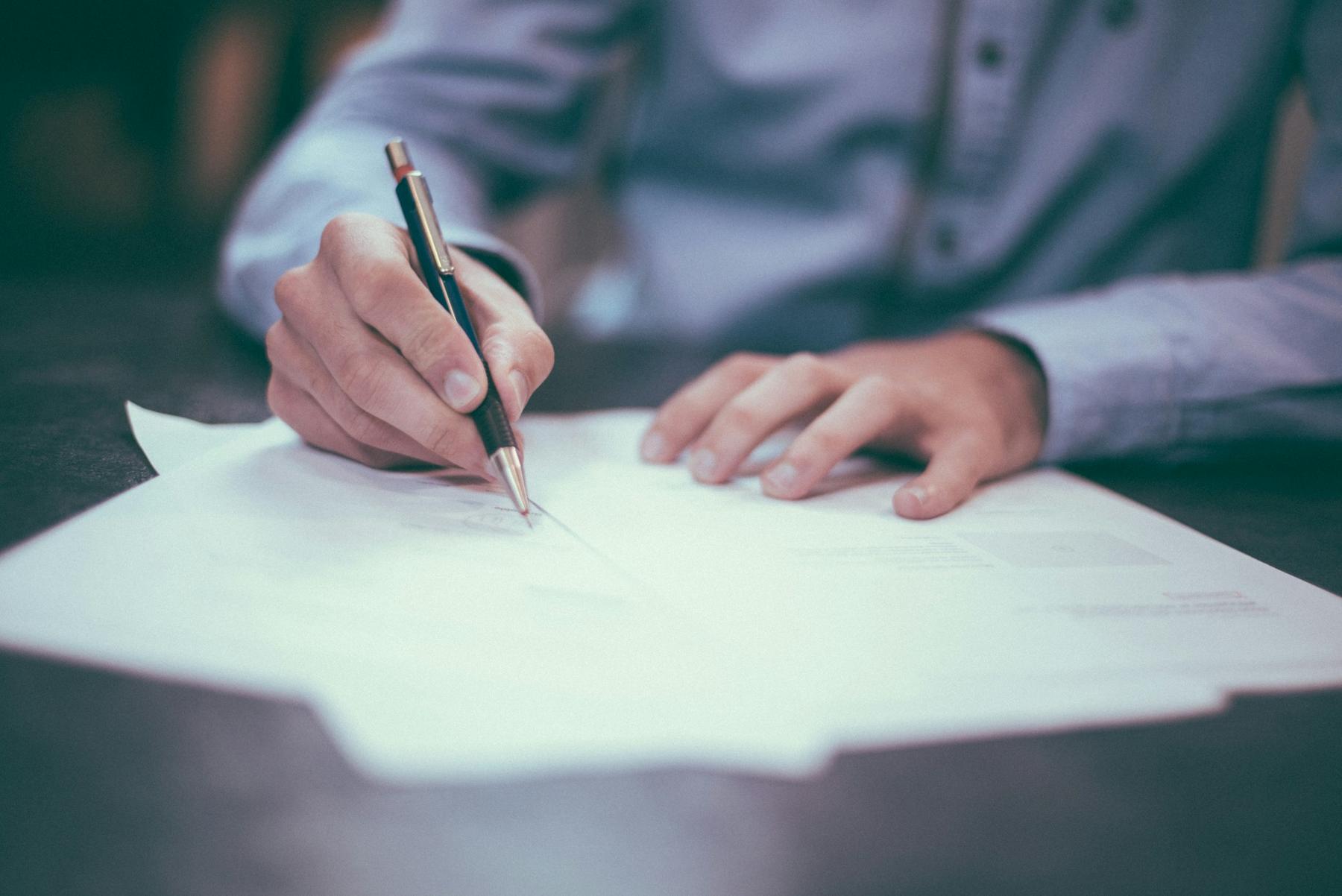 A person in a blue shirt writes on a piece of paper with a pen at a desk.