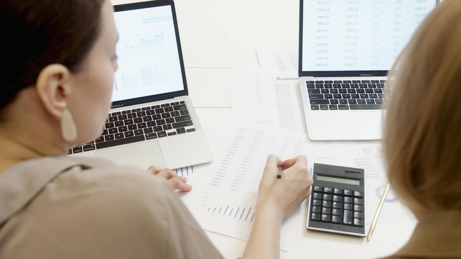 Two people work at a desk with laptops, financial documents, and a calculator, analyzing data and making notes.