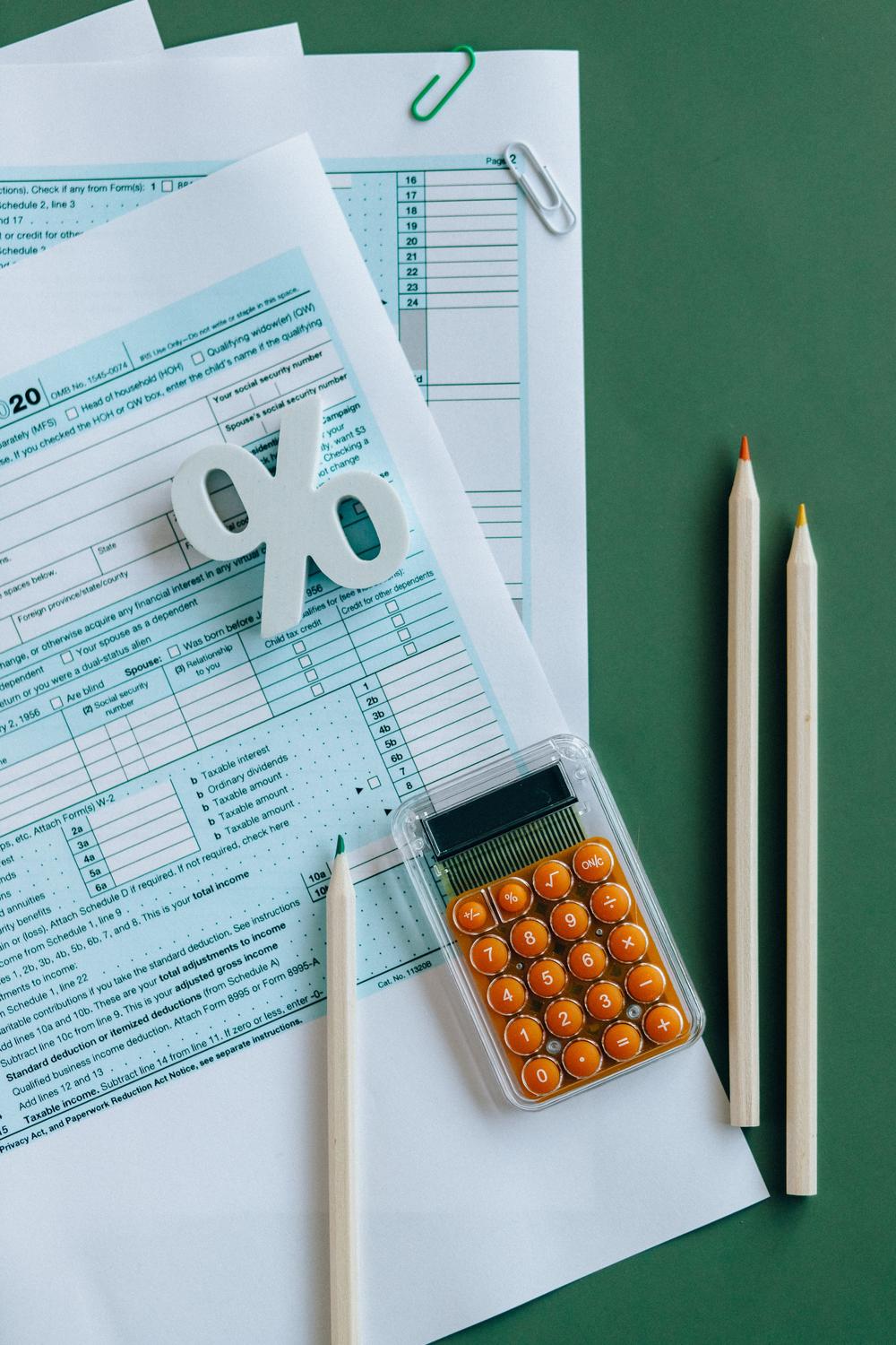 Tax documents, a calculator, a percentage symbol, and three pencils are arranged on a green surface.