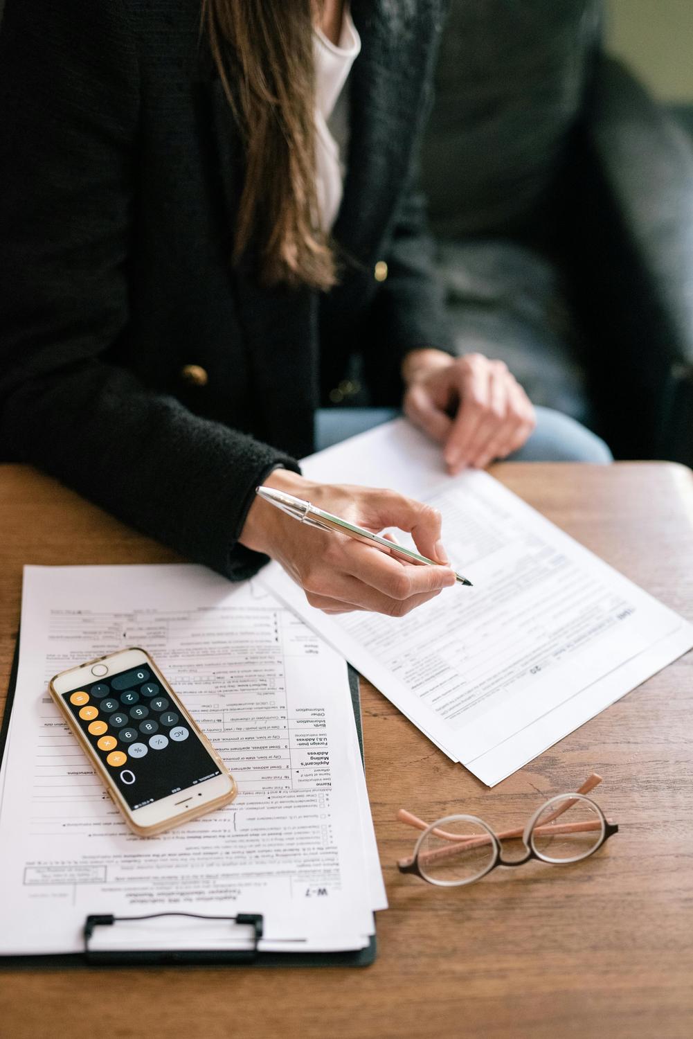 A person reviews and fills out paperwork at a desk with tax forms, a smartphone displaying a calculator app, and a pair of eyeglasses.