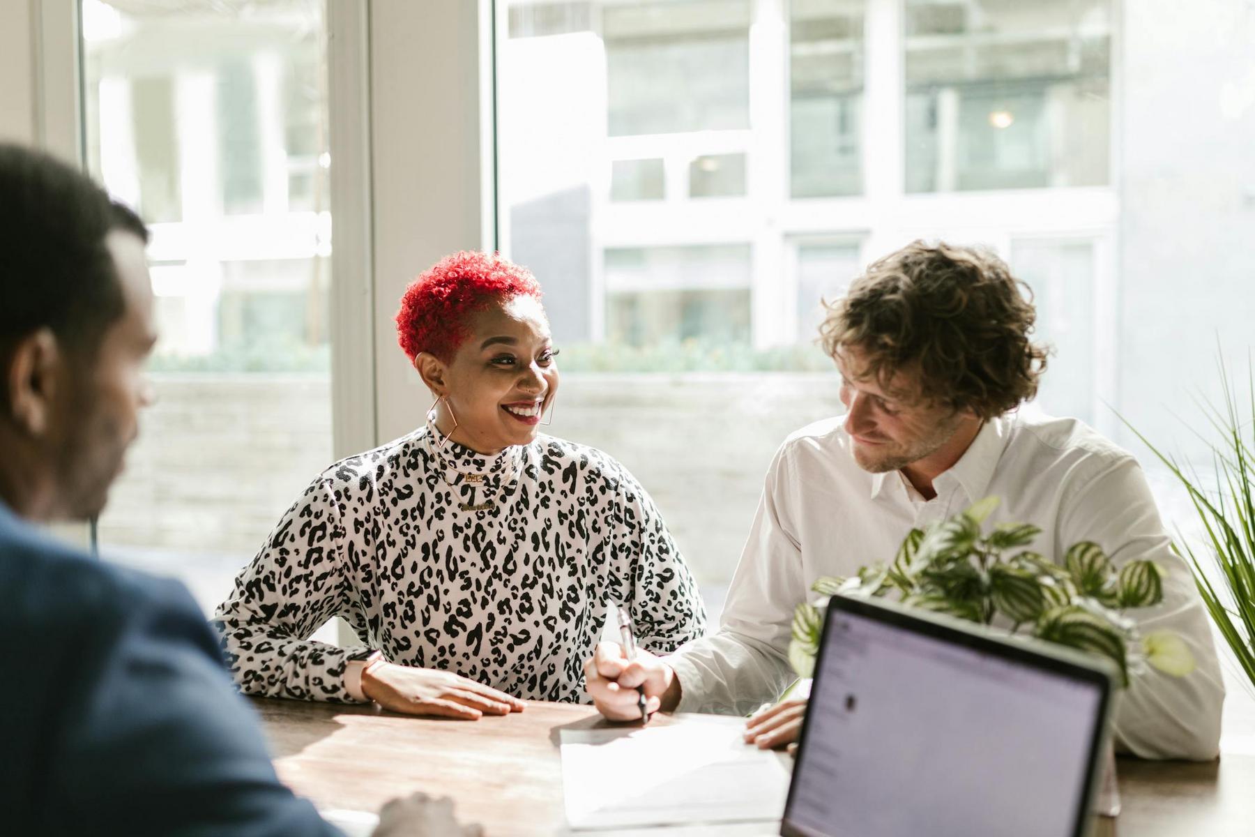 Three people sit at a table in a bright office, talking and smiling, with a laptop and some papers in front of them.