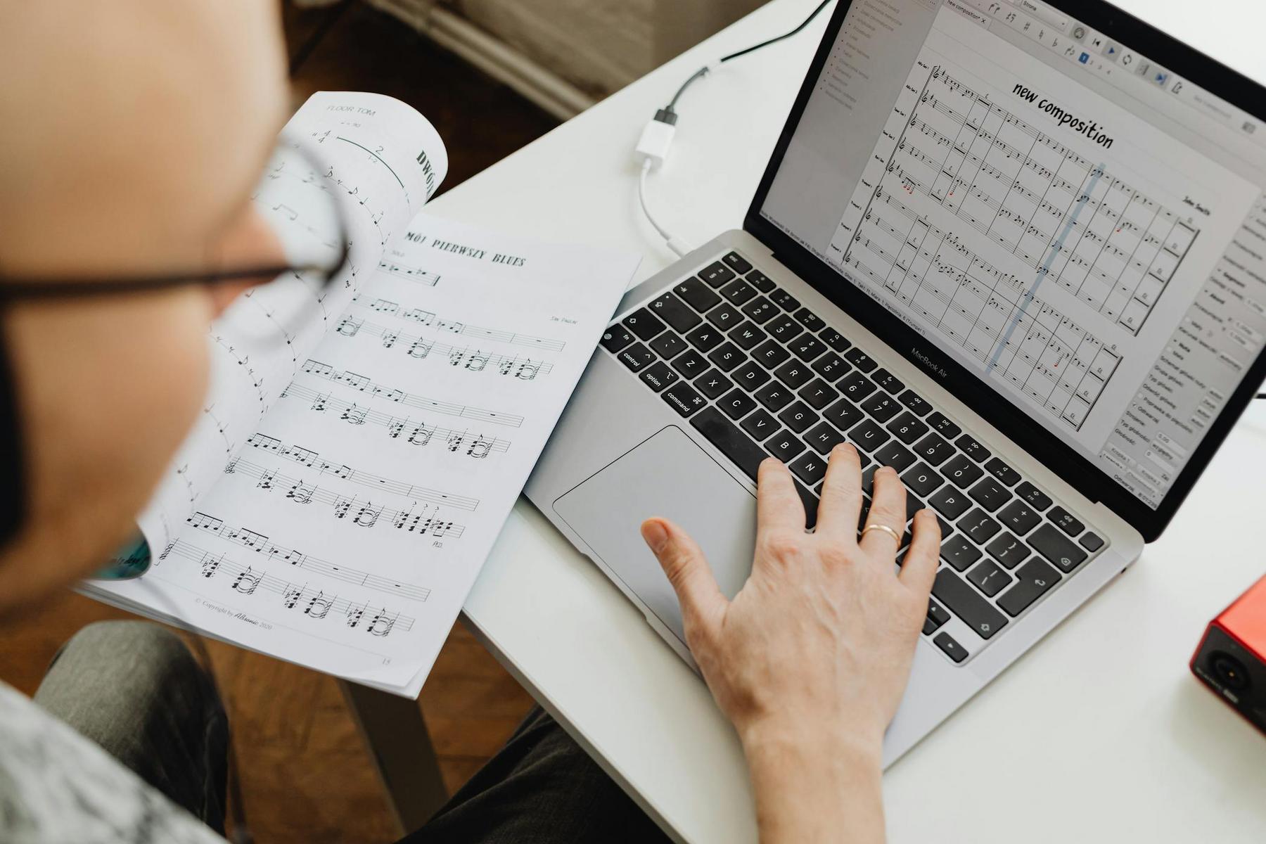 Person reviewing sheet music in a book while editing a musical composition on a laptop at a desk.