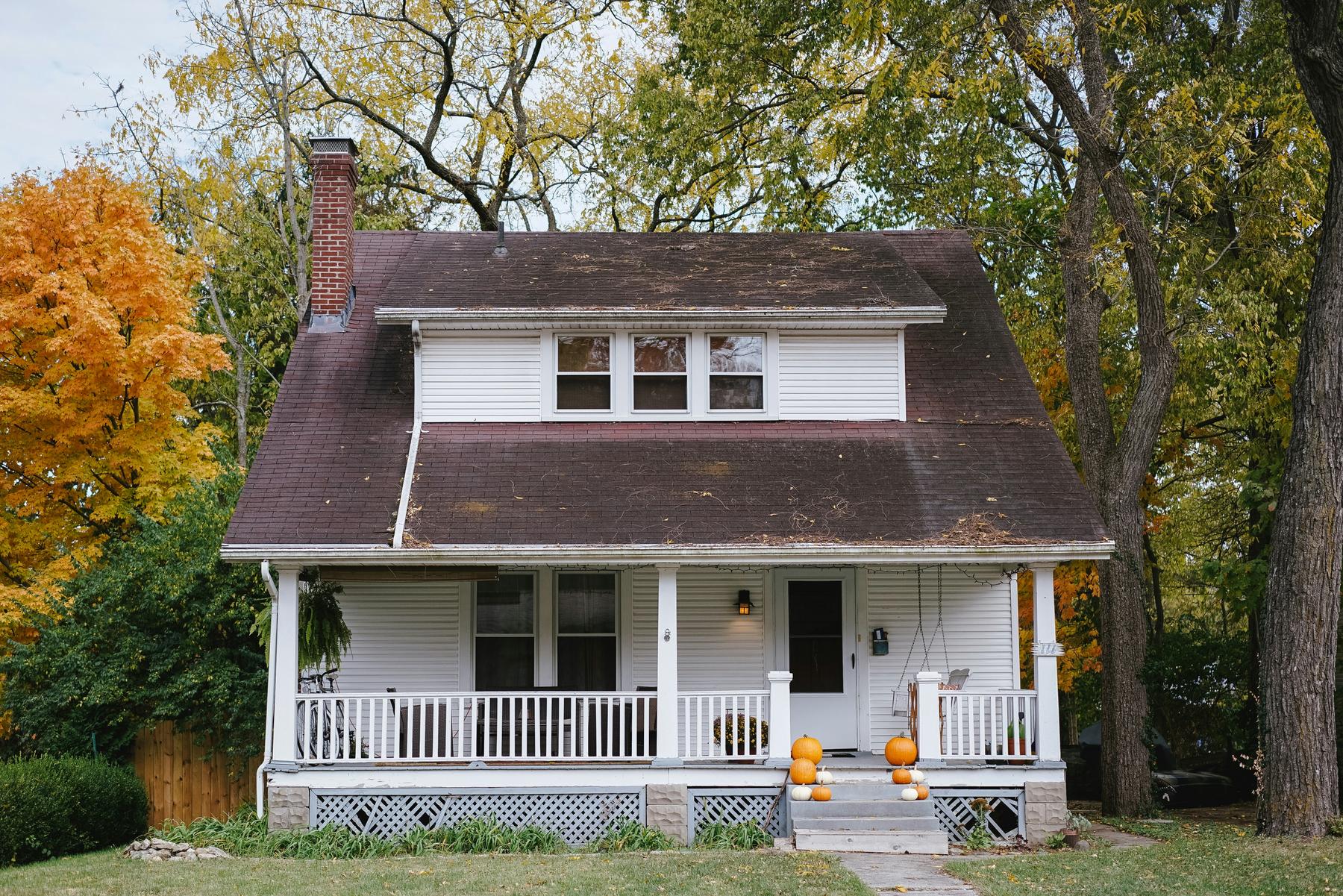 A two-story house with a porch, surrounded by autumn trees. Four pumpkins are lined up on the front steps.