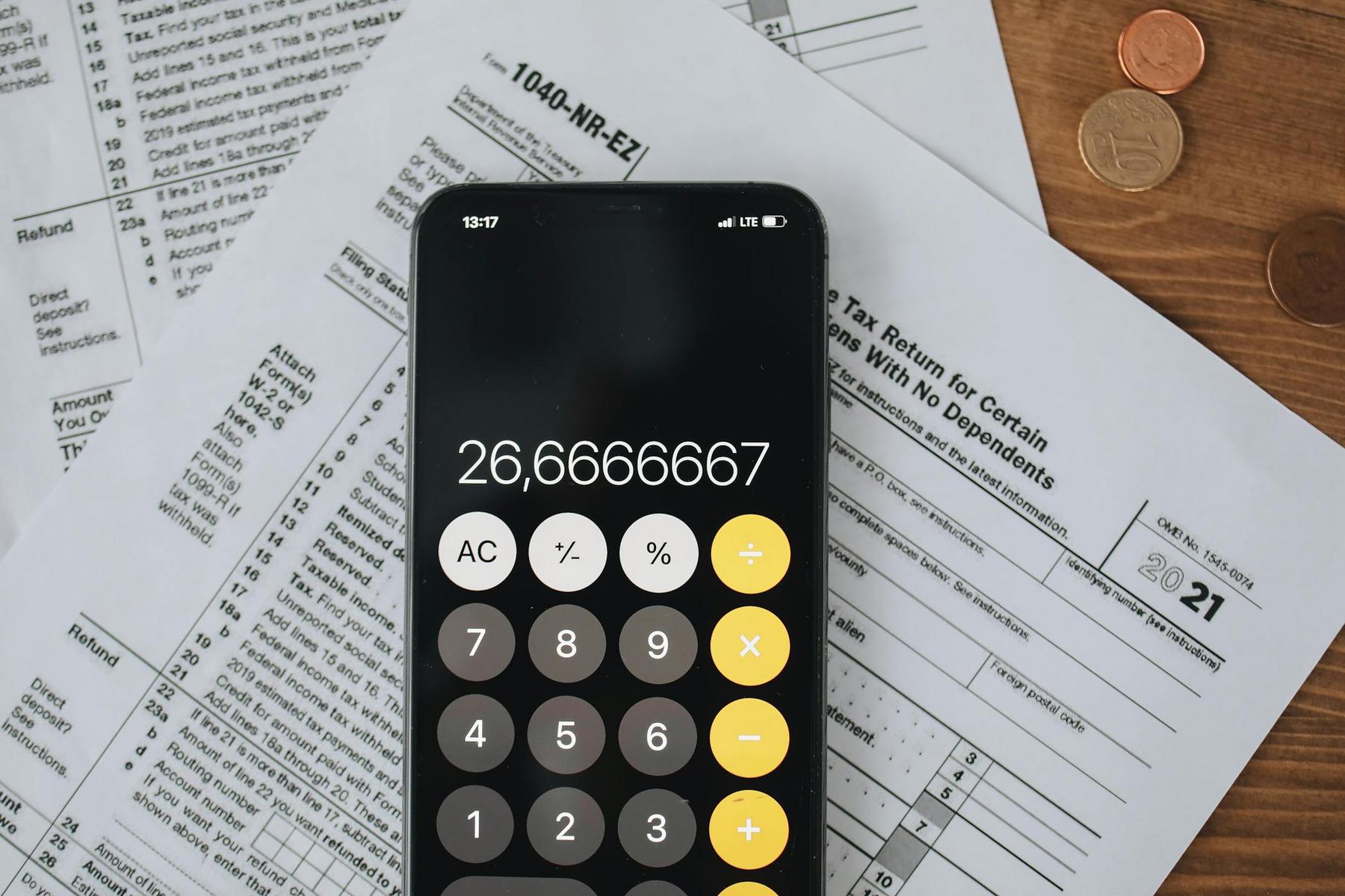 A smartphone displaying a calculator app sits on top of US tax forms, with a few coins scattered to the side on a wooden surface.