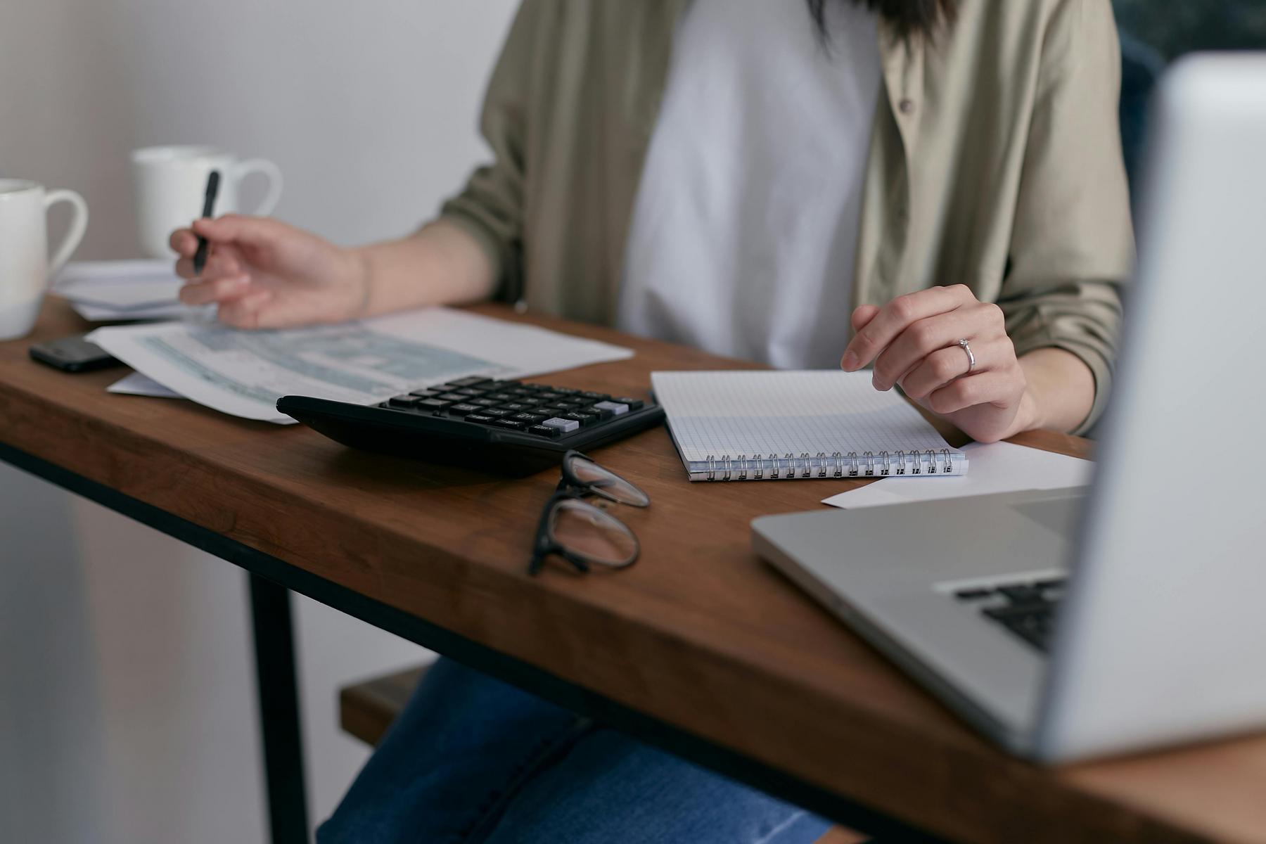 Person sitting at a desk working with papers, a calculator, a notepad, glasses, and an open laptop. Two mugs are also on the desk.